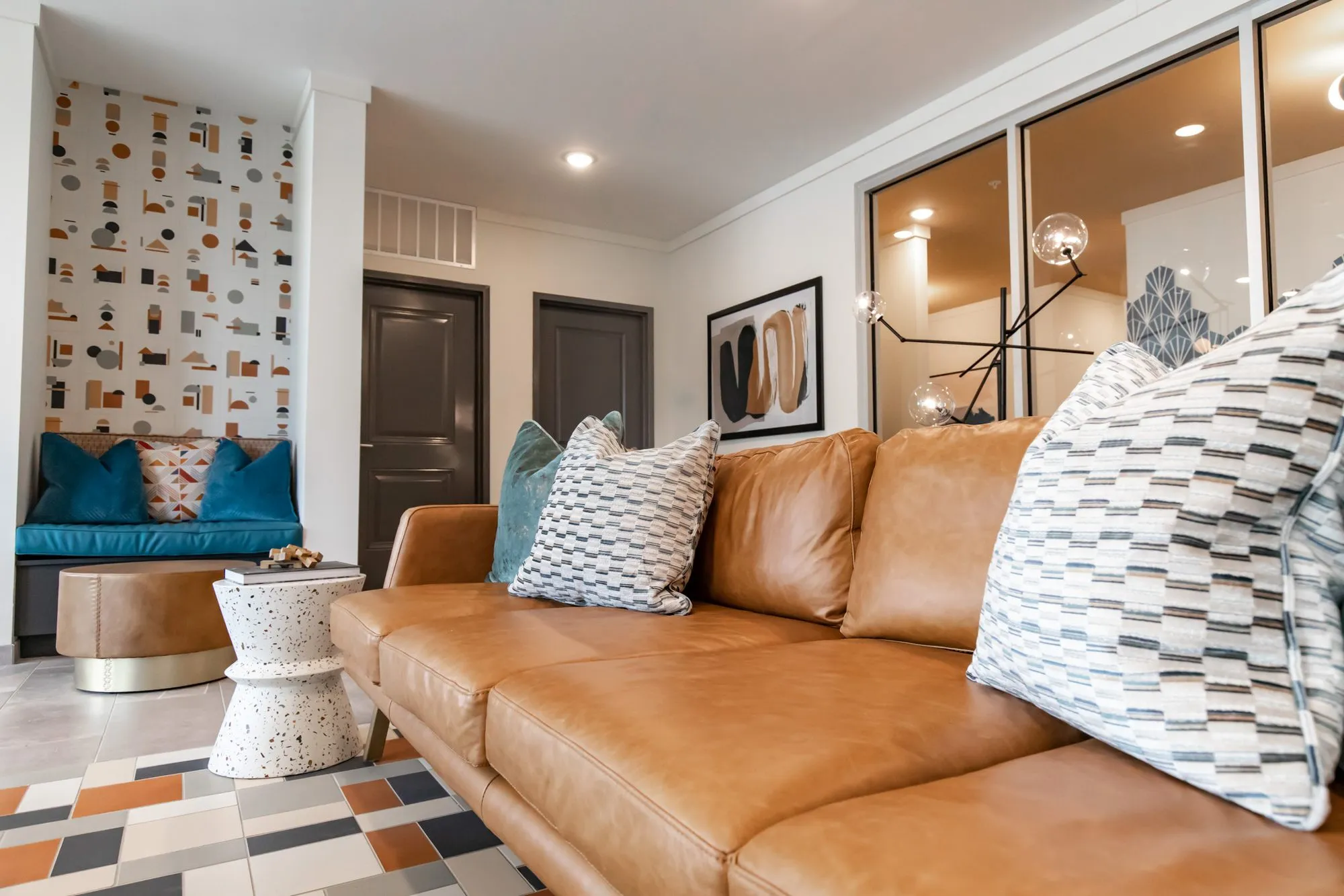 Multifamily amenity lobby lounge, a tan leather sofa with striped pillows toward a teal bench nook and terrazzo side table