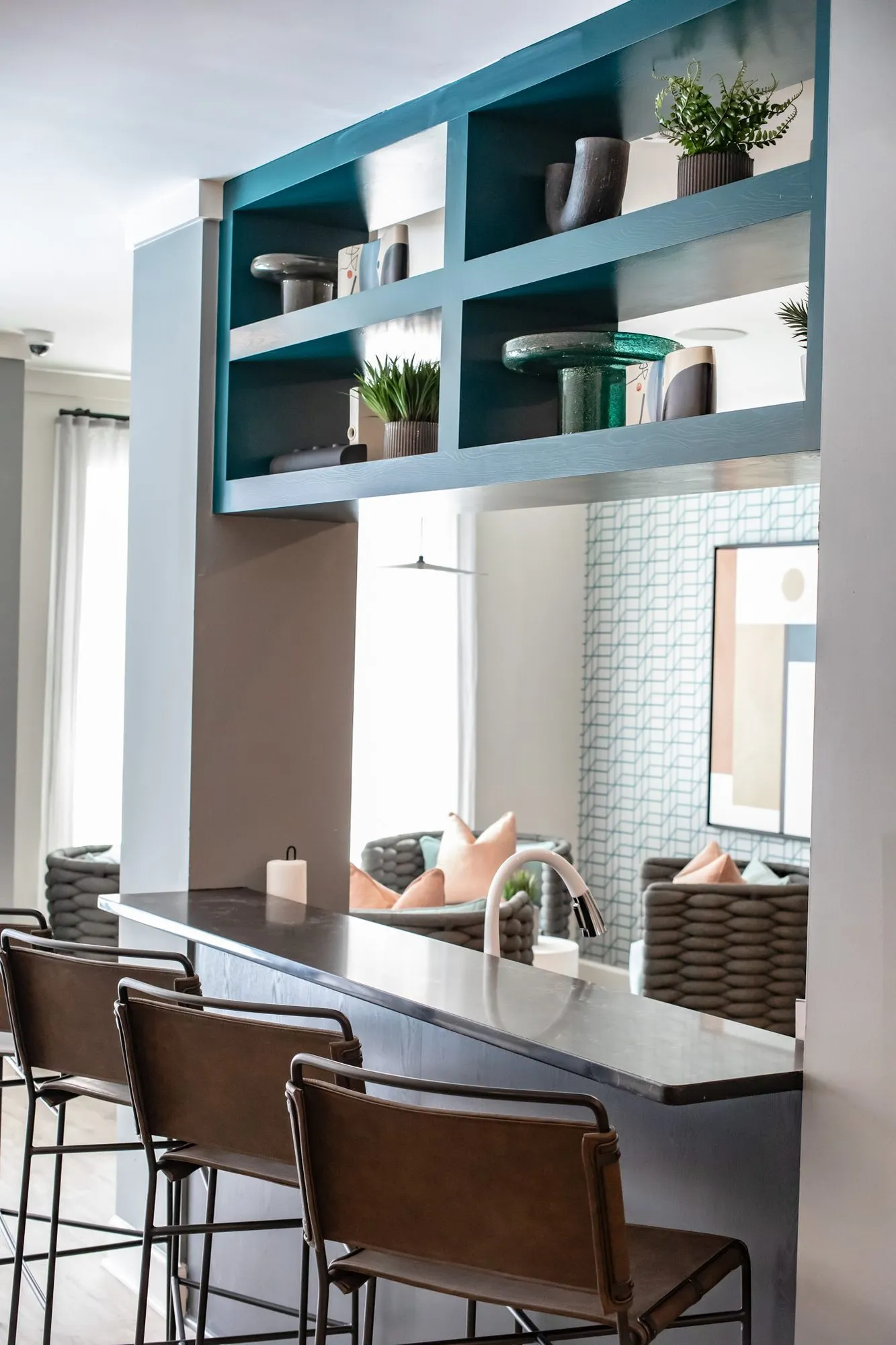 Multifamily amenity pass-through with teal open shelving above a dark bar counter and leather stools, lounge chairs beyond