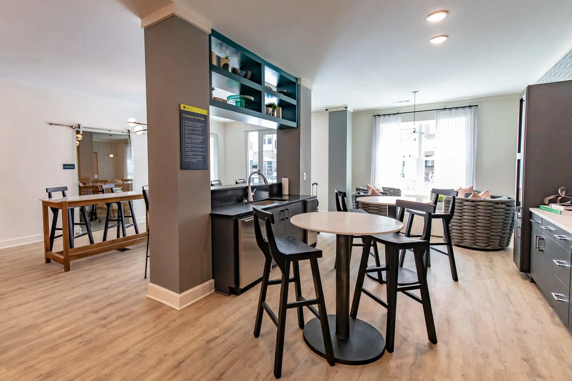 Multifamily amenity clubroom with tall black stools at round tables and a dark kitchenette under teal shelving