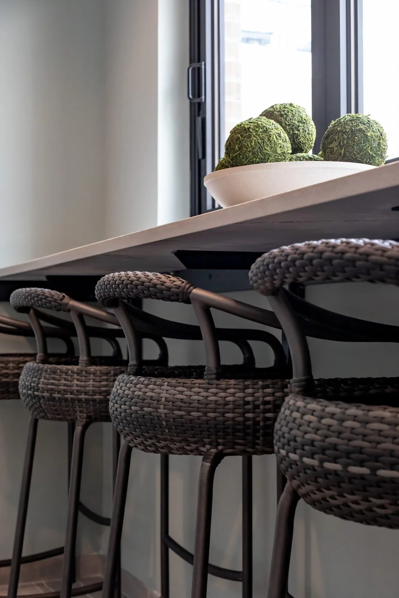 Multifamily amenity interior detail: woven brown bar stools at a dark counter with a bowl of moss balls in the window