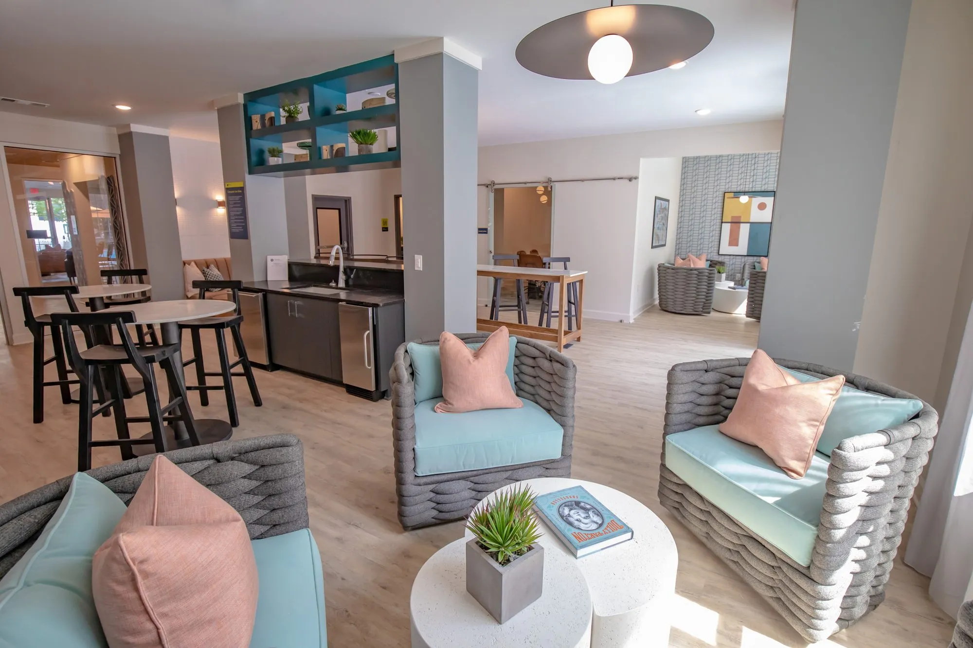 Multifamily amenity lounge of woven charcoal chairs around white tables, bar seating and teal shelves beyond