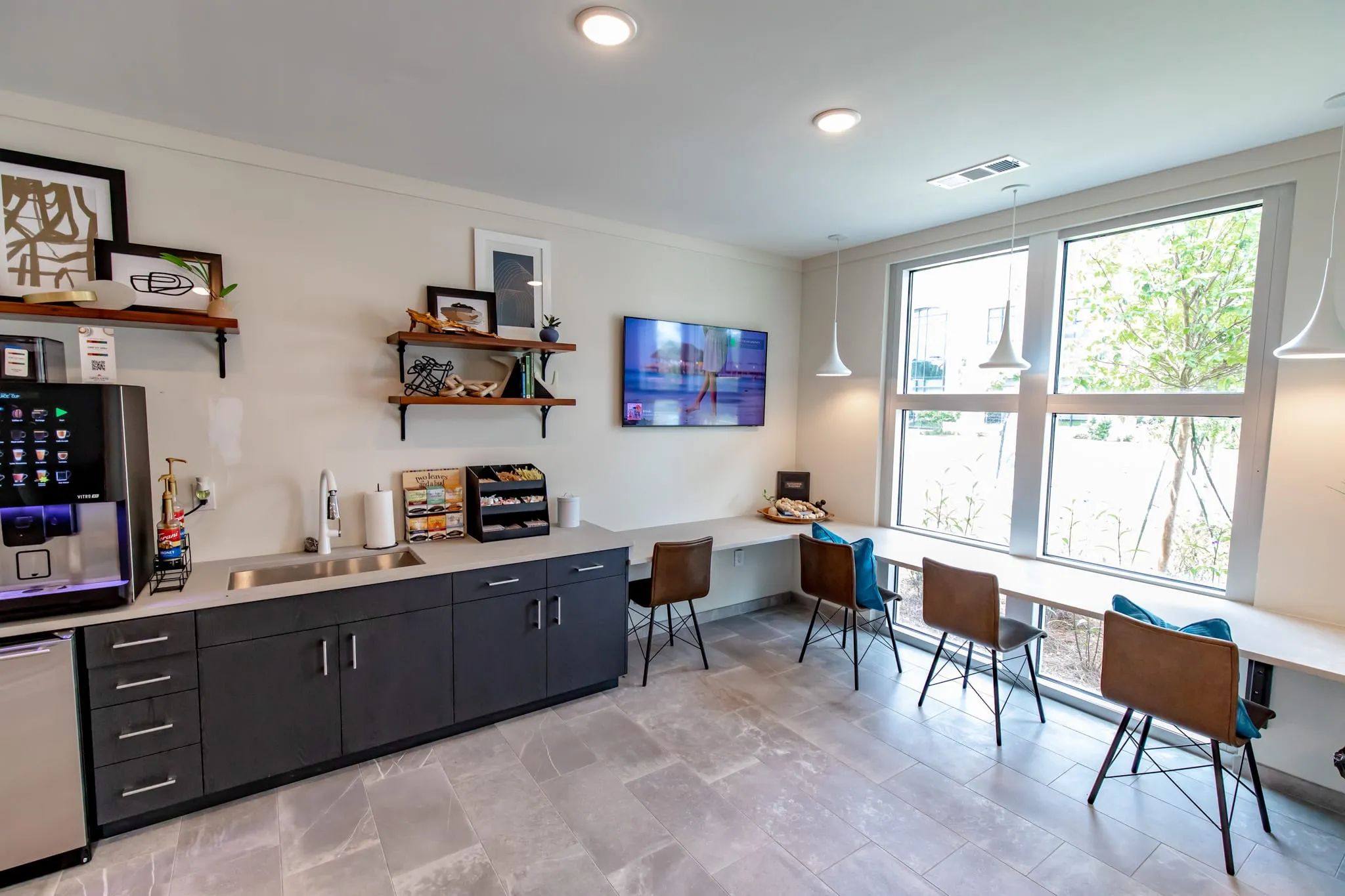 Multifamily amenity coffee bar with dark cabinetry and a window counter with leather stools below a wall-mounted screen