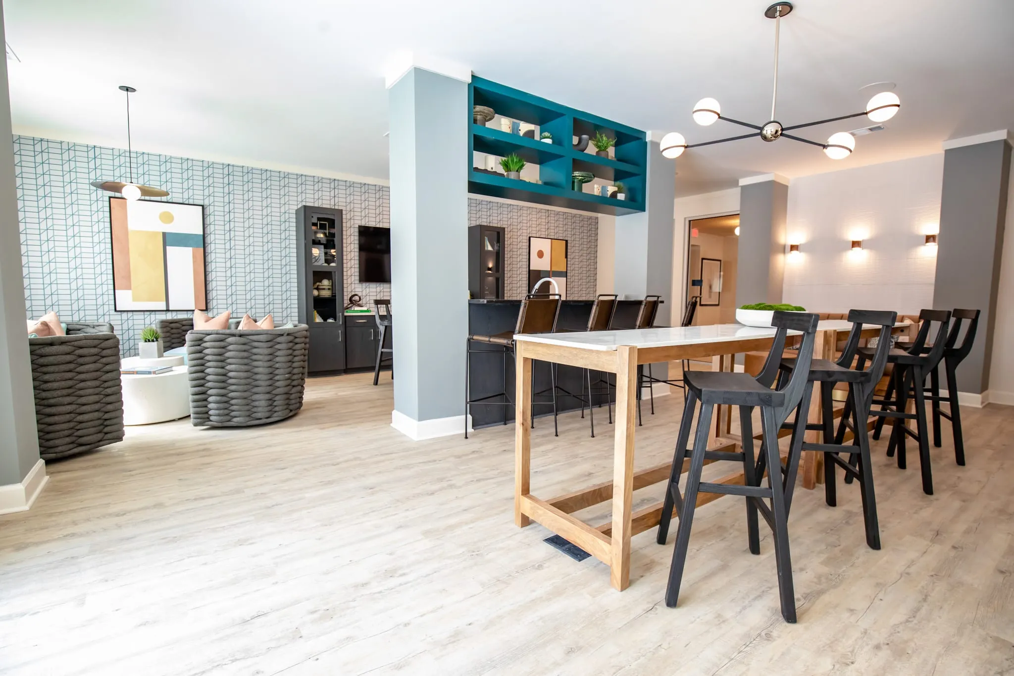 Multifamily amenity clubroom with a tall communal table and black stools beside teal shelving, woven lounge chairs beyond