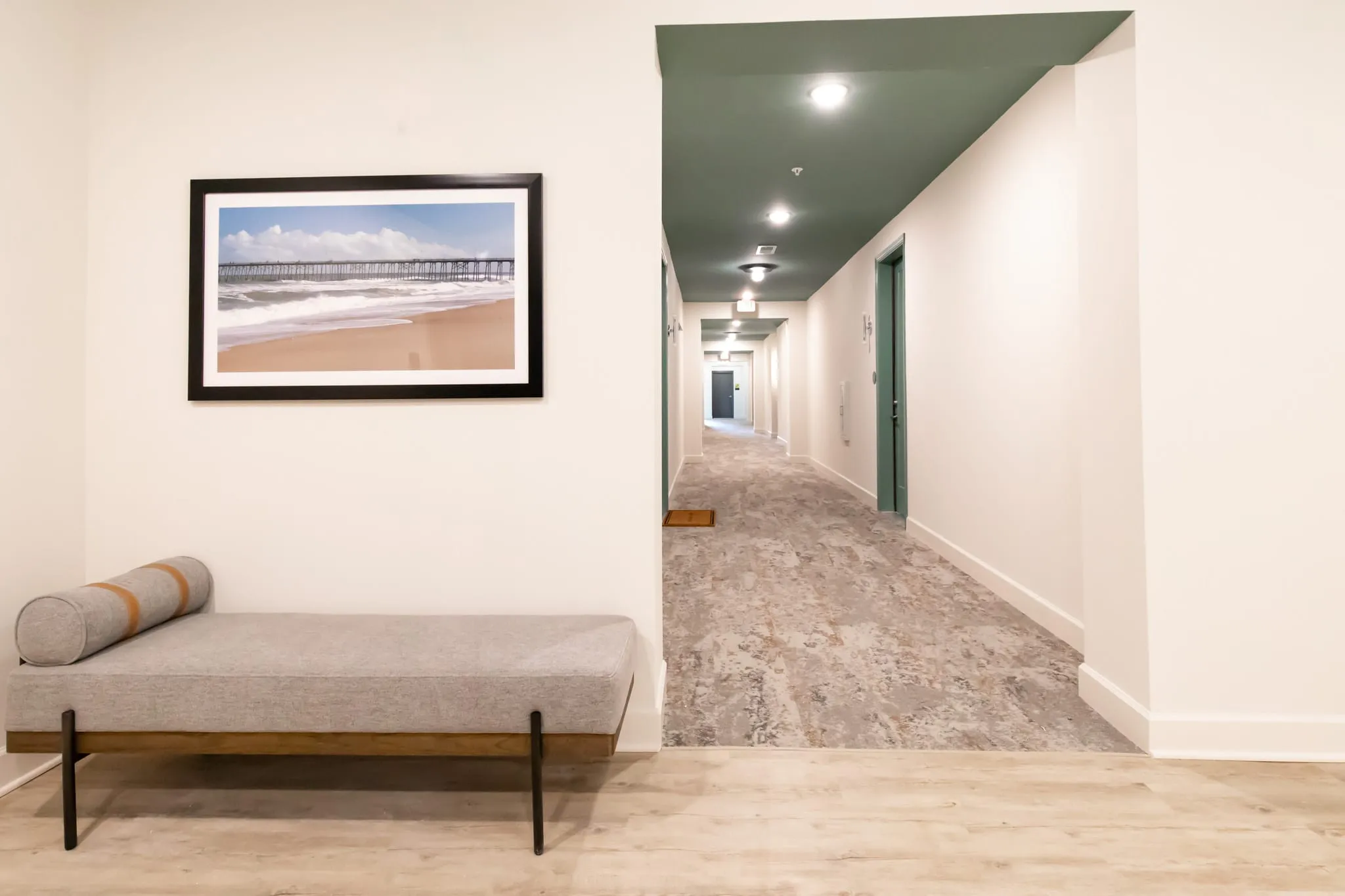 Multifamily residential corridor entry with an upholstered bench below a framed beach photograph, a green ceiling beyond