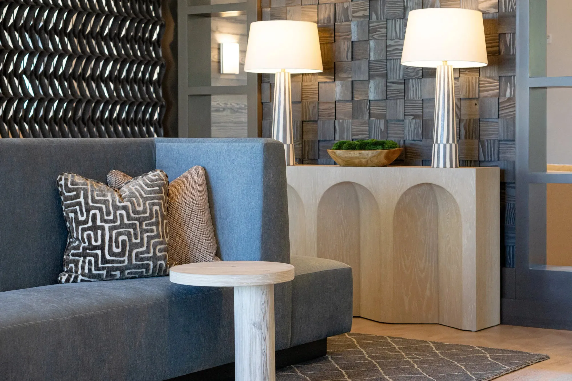 Multifamily amenity lobby seating with a grey banquette, a pale wood arched console, two tall silver lamps and a sculpted wood wall panel