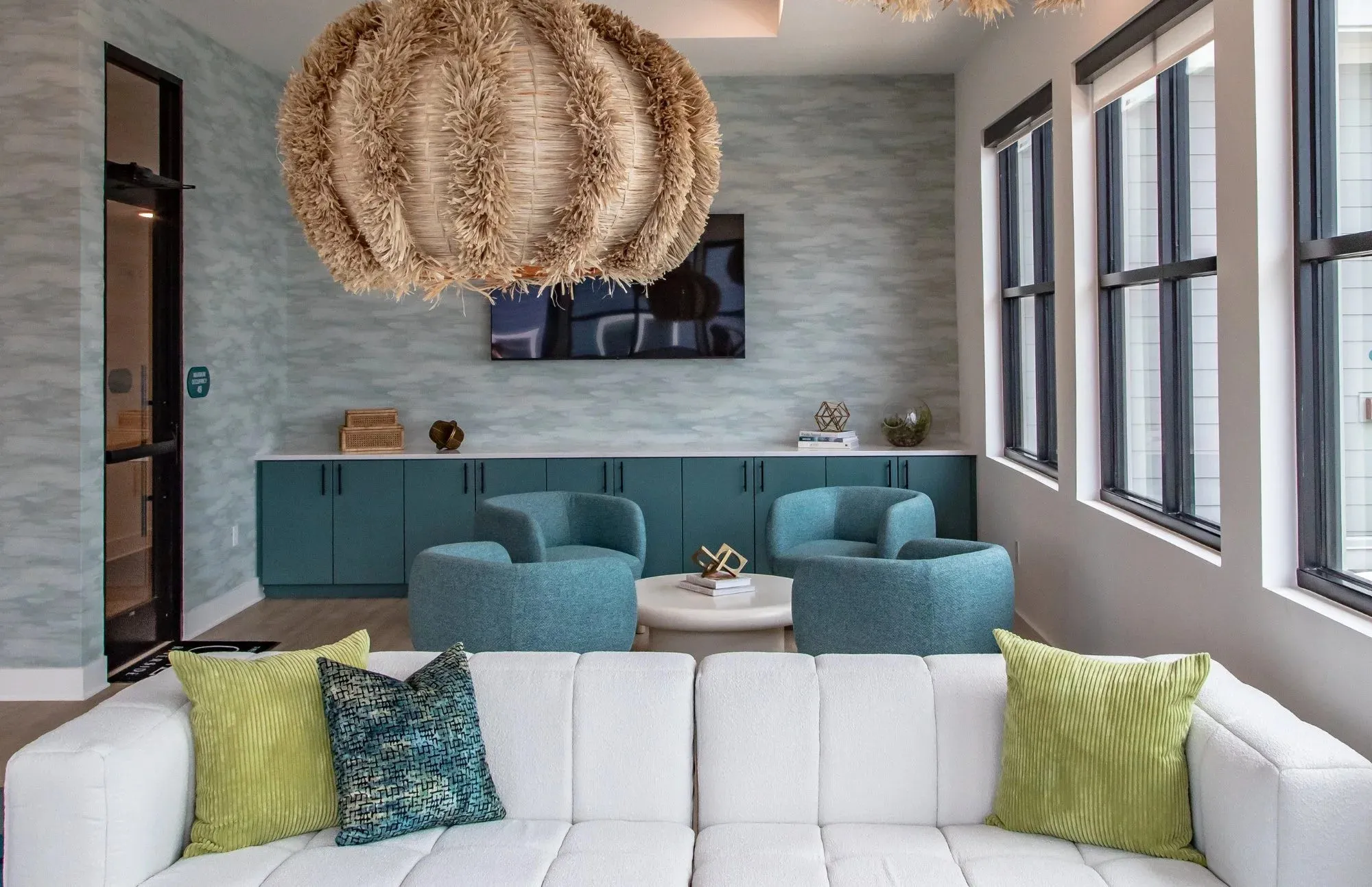 Lounge with an oversized fringed rattan pendant, two teal barrel chairs, teal built-in cabinetry and a white tufted sofa with green pillows