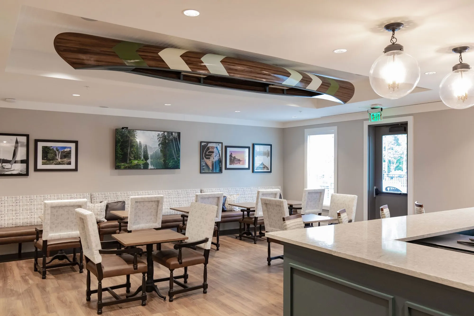 Senior living bistro seating under a ceiling mounted canoe, cream upholstered chairs at small tables and framed photographs