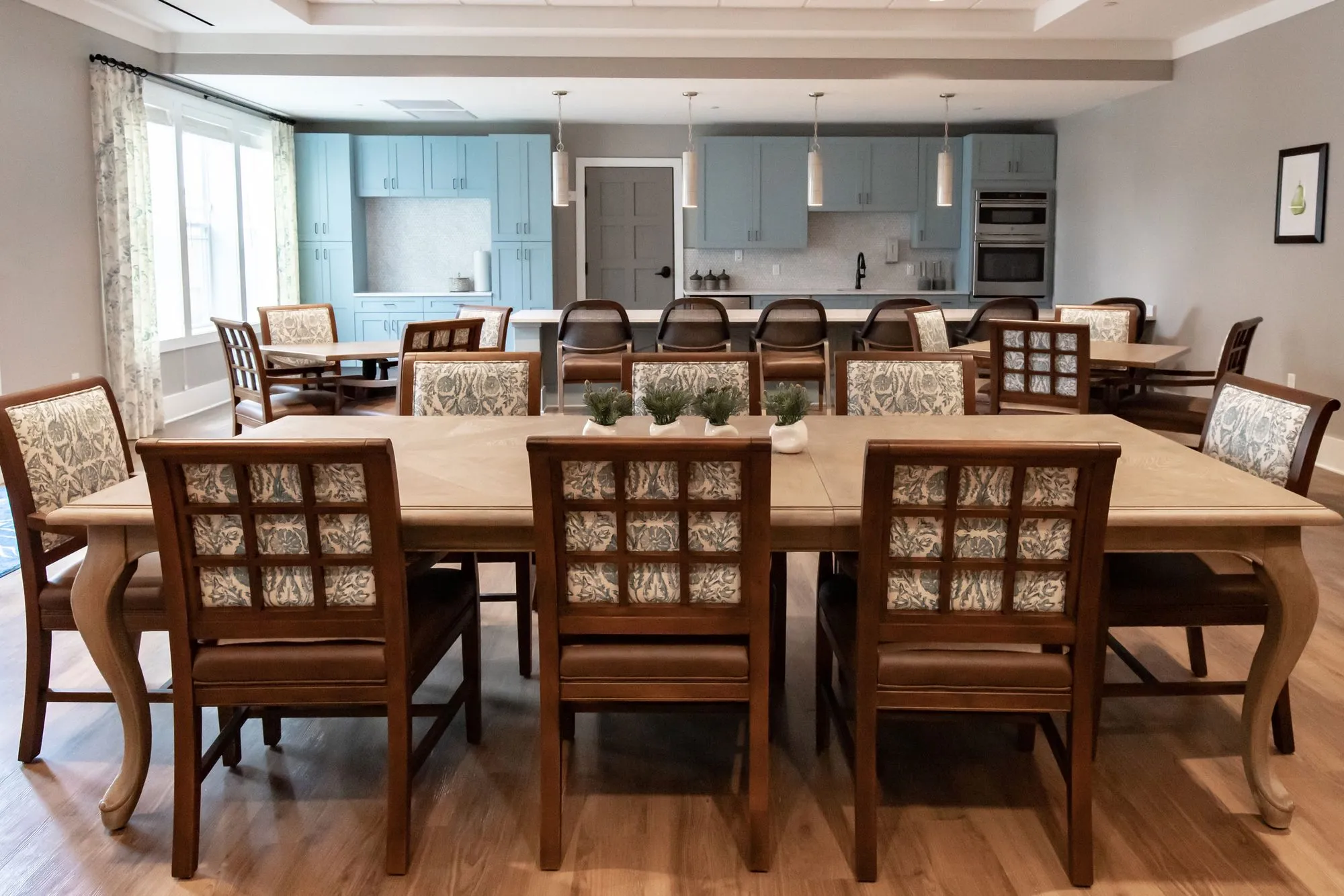 Senior living activity room with a long wood table and lattice back chairs, blue cabinet kitchen and barstools behind