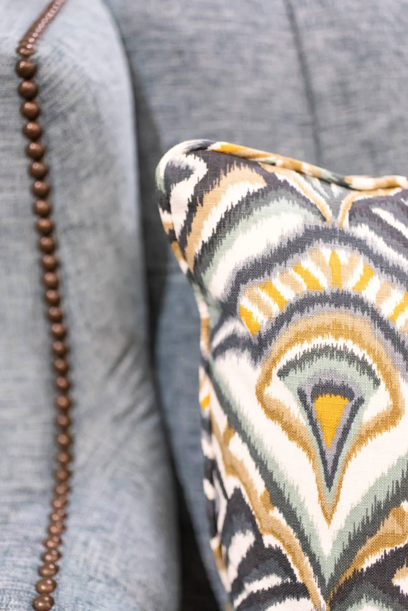 Senior living interior detail: nailhead trim on a grey sofa beside an ikat feather pattern pillow in gold, grey and green