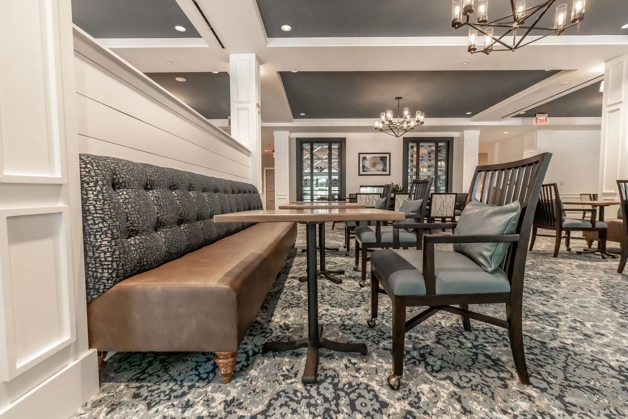Senior living dining room with a tufted banquette, slat back chairs and a patterned blue carpet under navy ceiling coffers