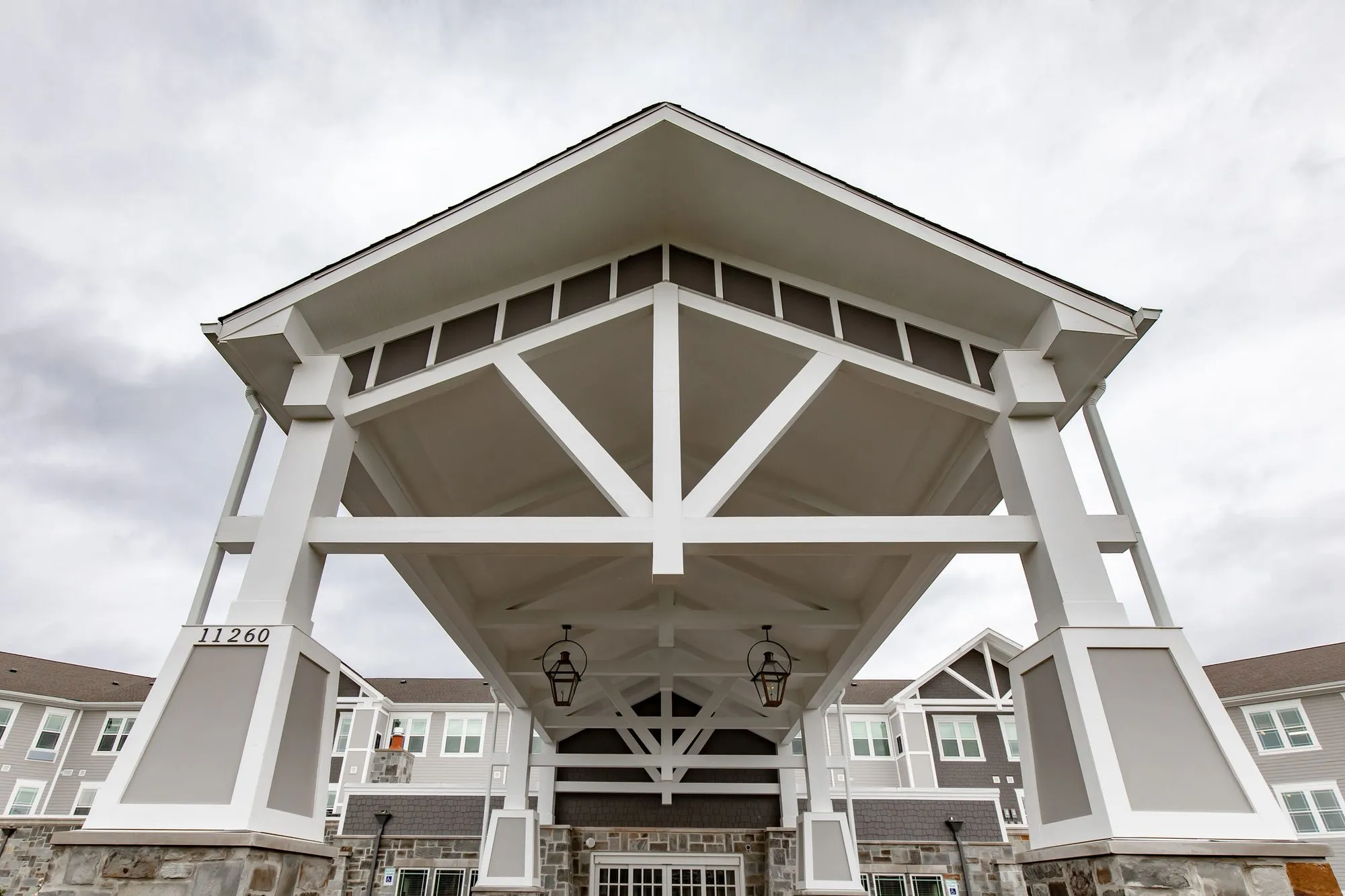 Senior living community exterior: porte cochere with white timber trusses and lanterns before the stone and sided entrance