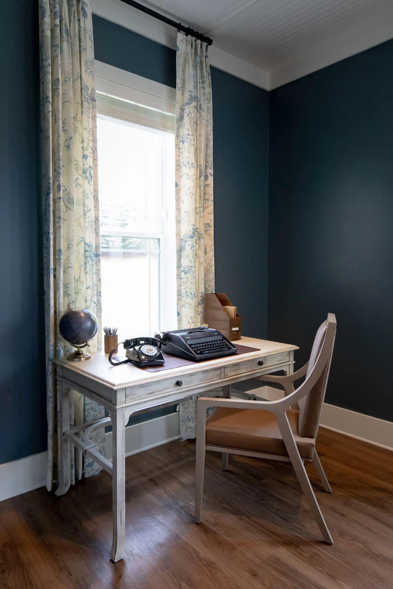 Senior living writing nook in a navy room, a whitewashed desk with a typewriter and chair below a floral curtained window