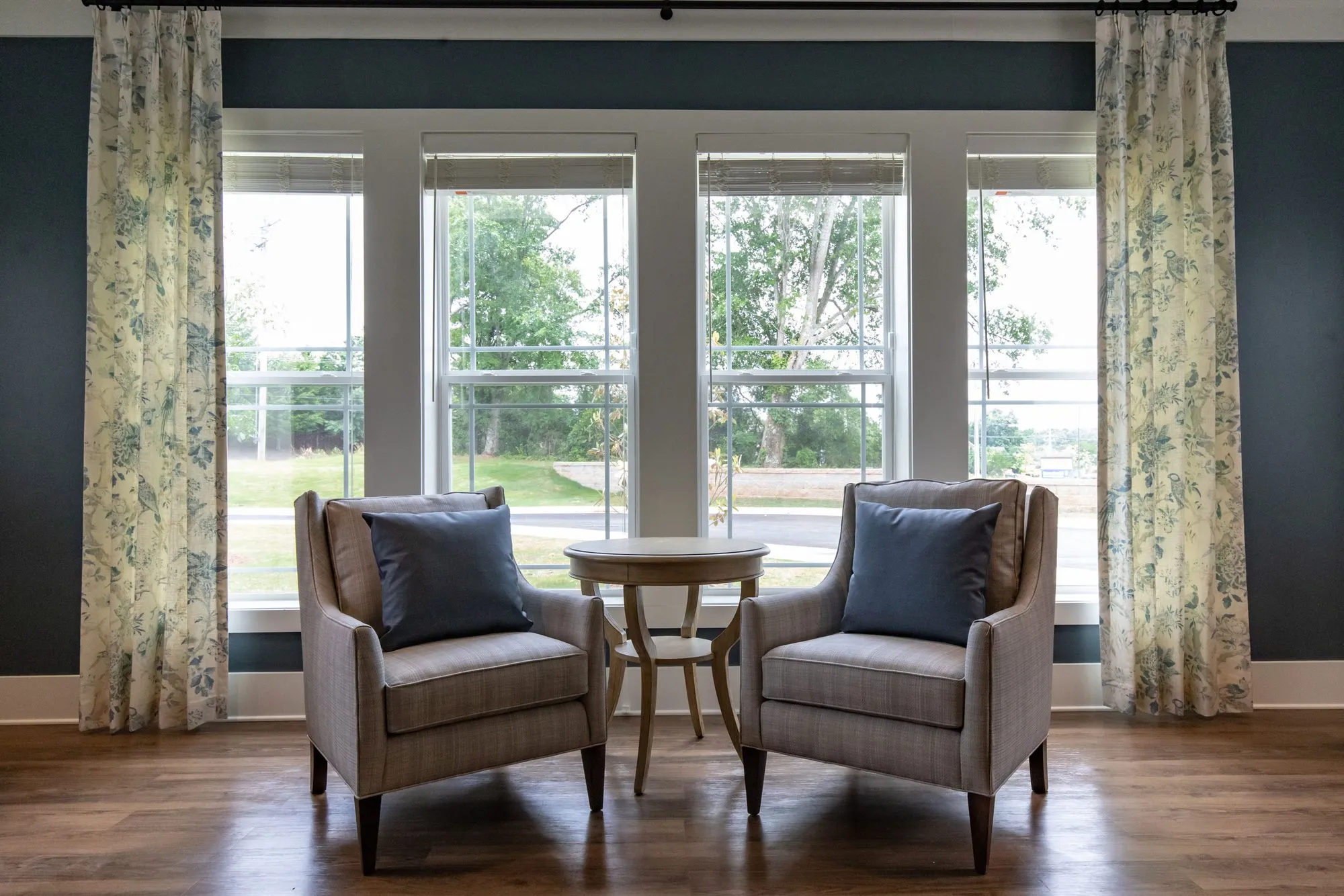 Senior living interior detail: two plaid armchairs and a round side table before a floral curtained window on a navy wall