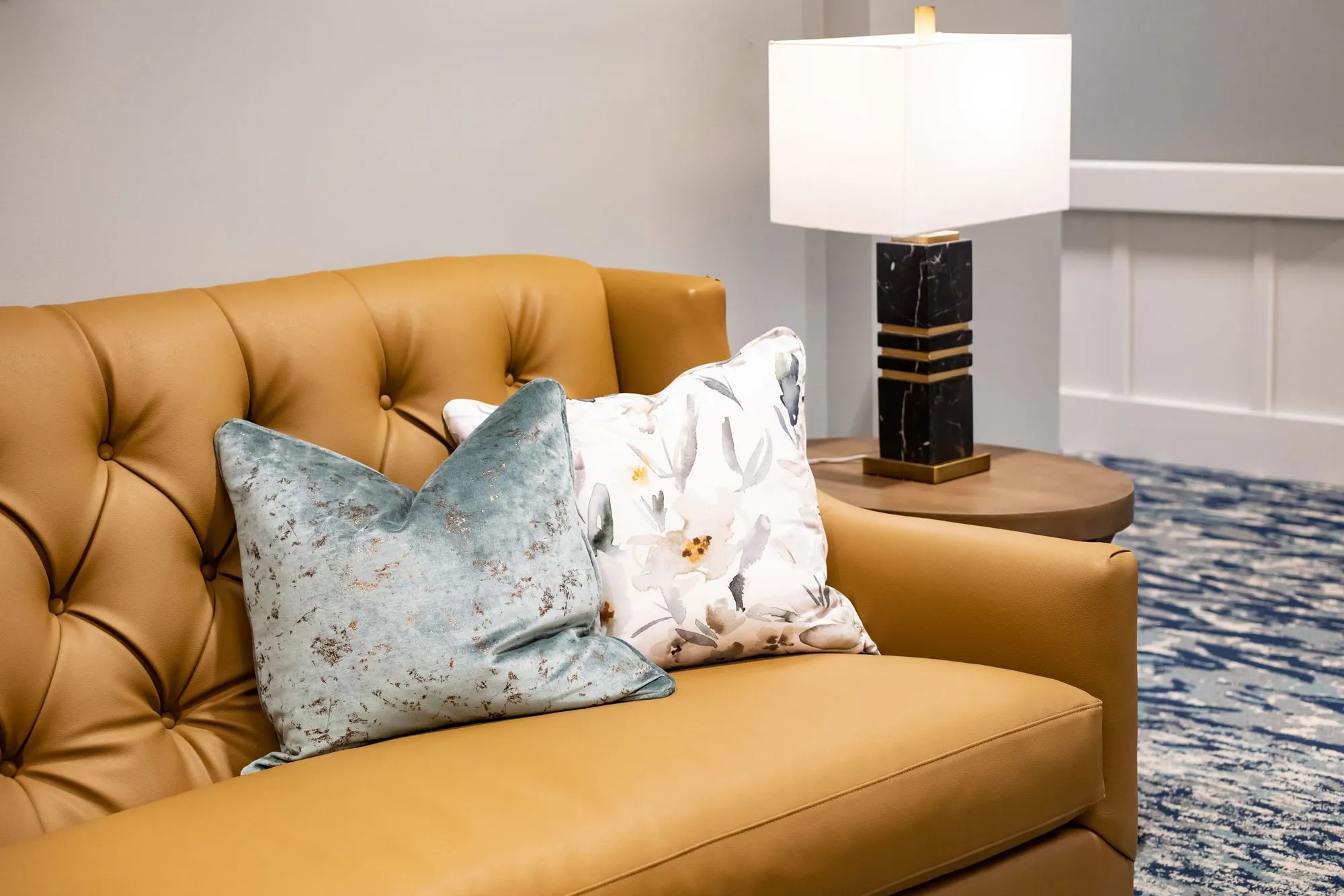 Senior living interior detail: camel tufted leather sofa with blue and floral pillows beside a black and brass lamp