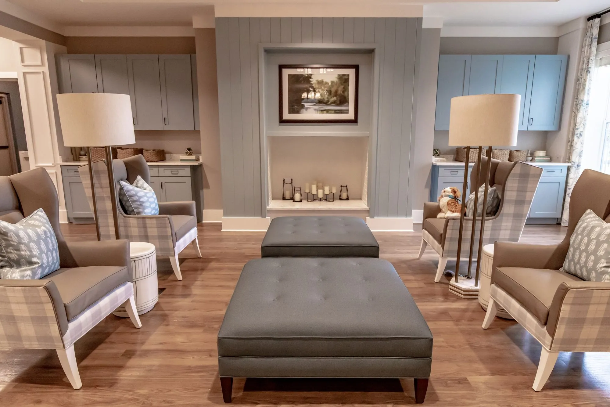 Senior living sitting room with plaid wing chairs, grey tufted ottomans and floor lamps before a candle niche and cabinets
