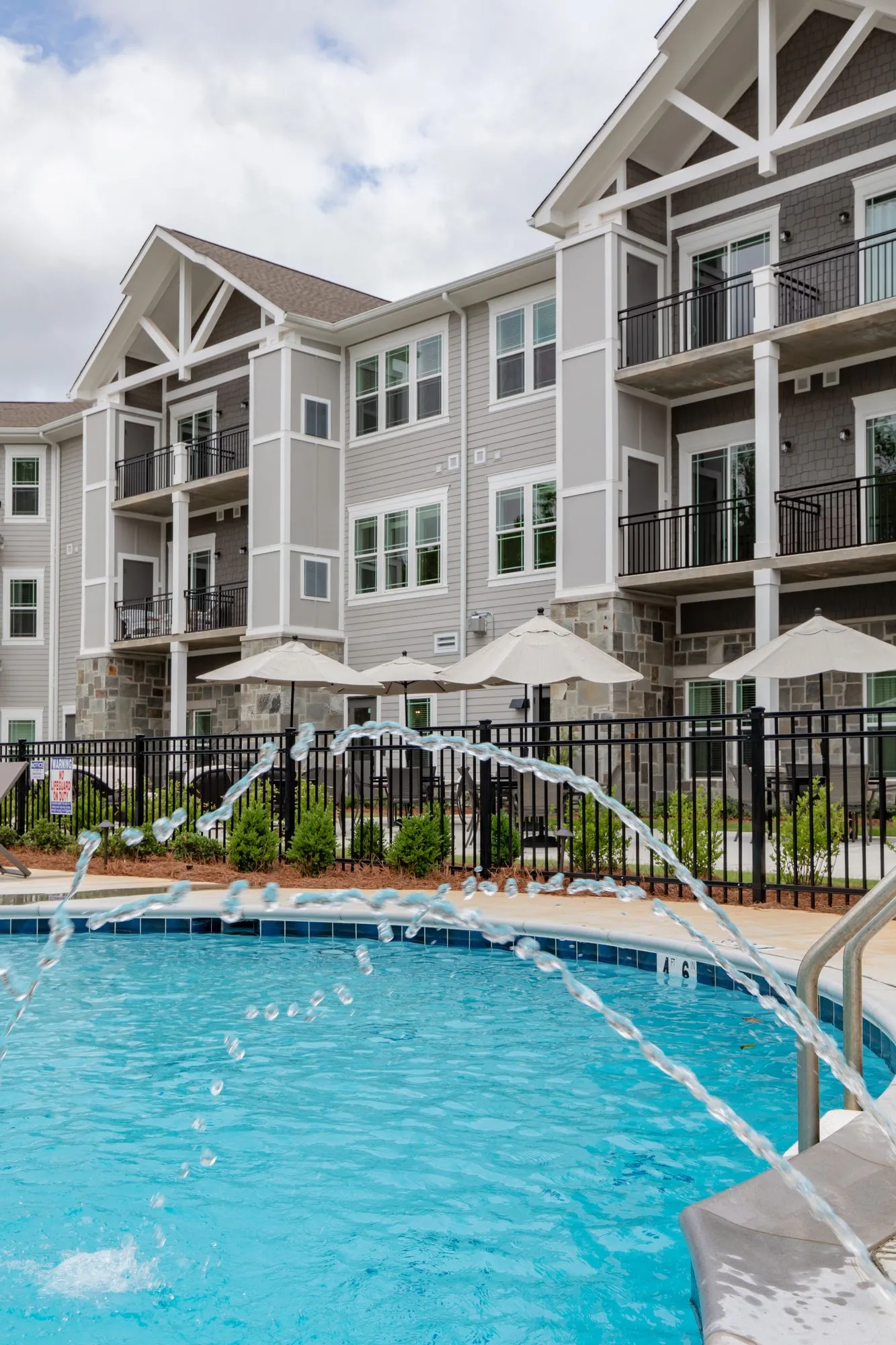 Senior living pool with fountain jets, umbrellas on the fenced patio and grey sided residences above