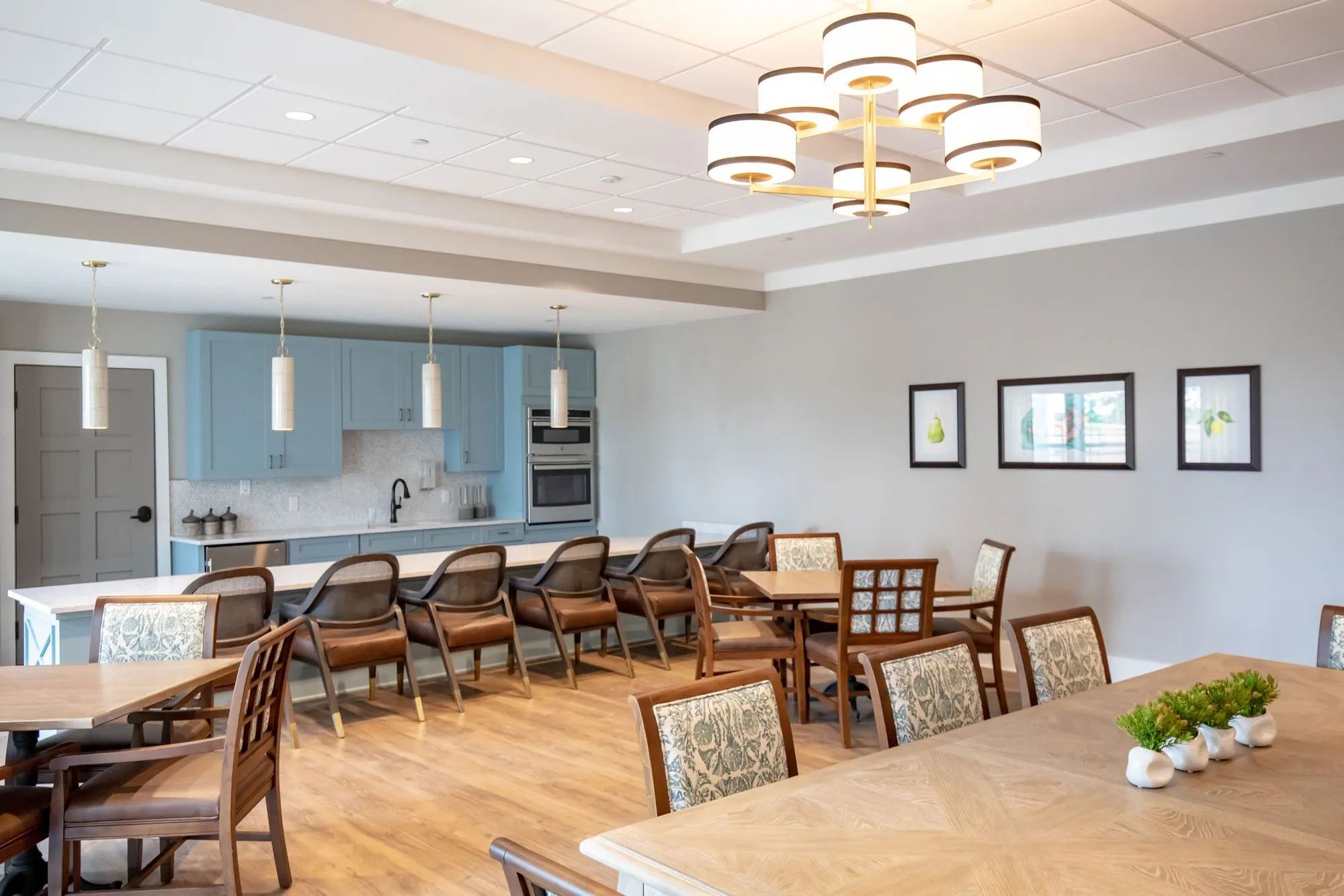 Senior living activity room under a tiered drum chandelier, wood tables and chairs, blue kitchen cabinets at back