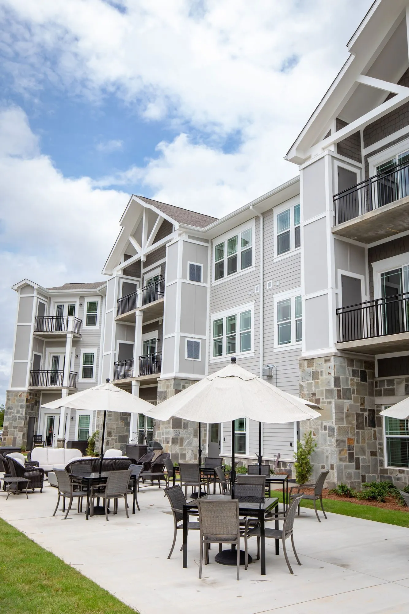 Senior living patio with white umbrellas over dark wicker dining sets at the foot of stone and grey sided residences