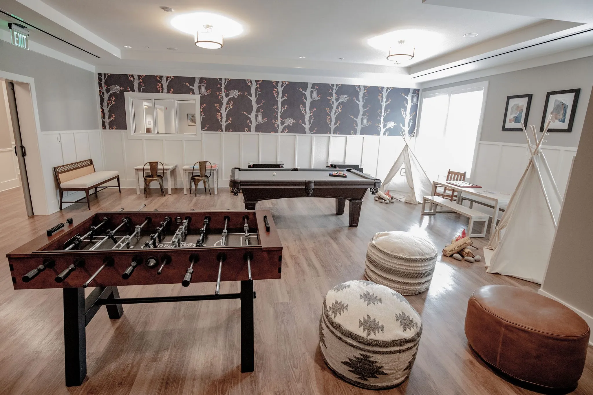 Senior living game room with foosball and pool tables on wood flooring below tree-print wallpaper