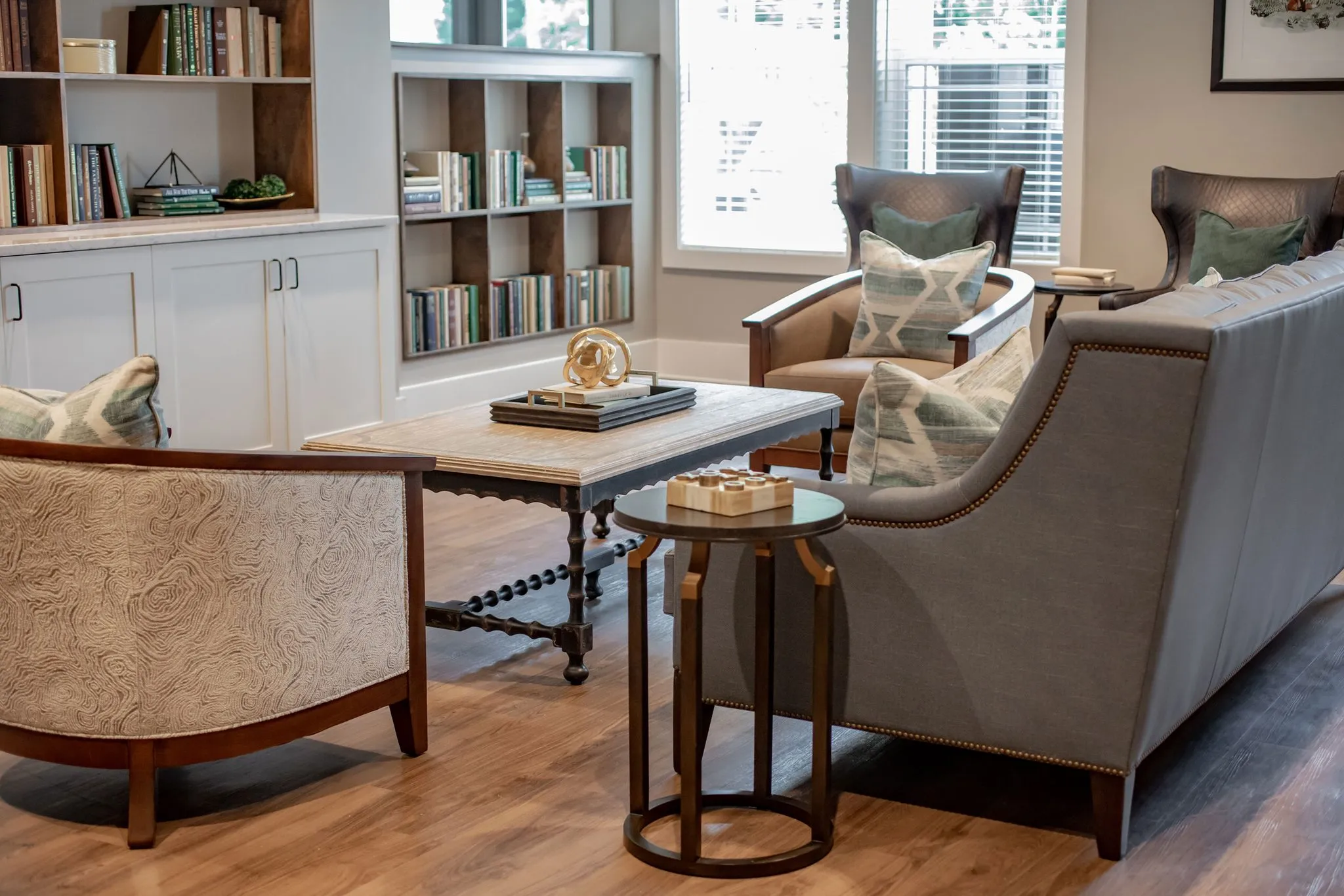 Senior living library sitting area with built-in bookshelves, a nailhead sofa, wing chairs and a turned-leg wood table