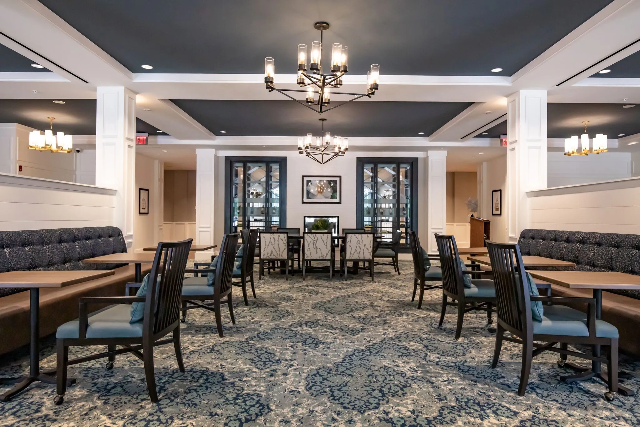 Senior living main dining room with brass chandeliers under a navy coffered ceiling above navy banquettes and a floral carpet