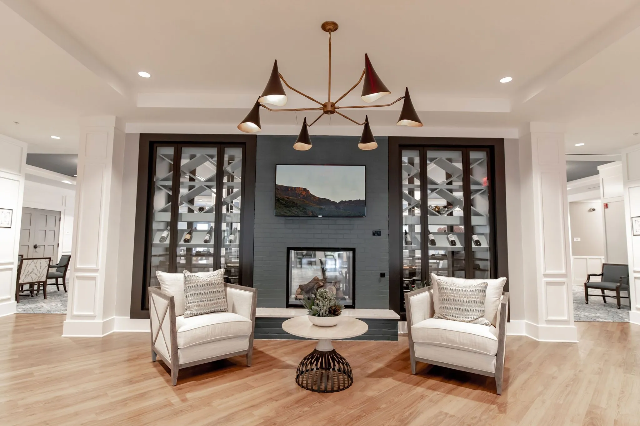 Senior living lounge with dark wine racks flanking a TV and fireplace, white armchairs below a cone pendant chandelier
