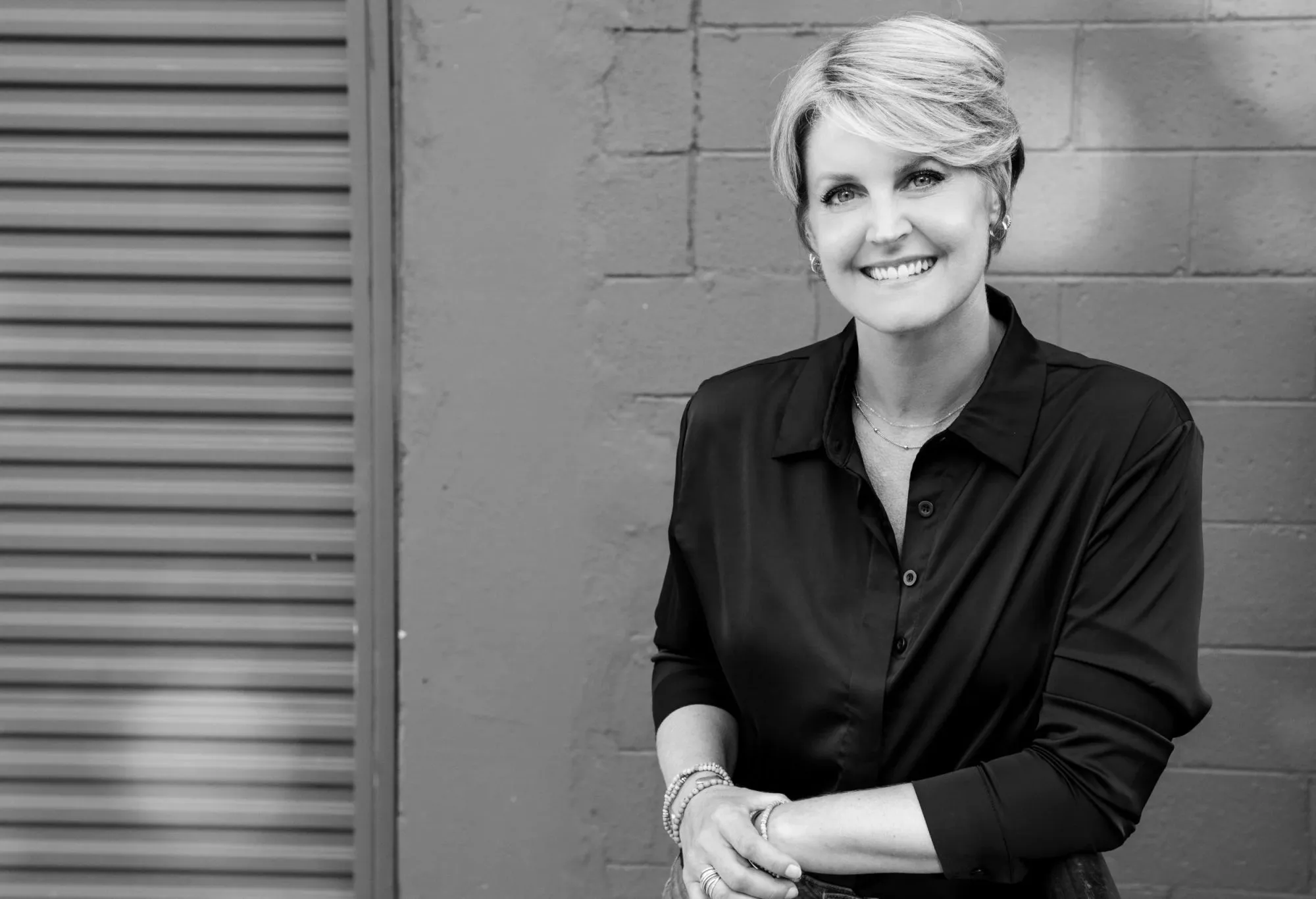 Melissa Banko in black and white, leaning against a painted brick wall beside a rolling steel door