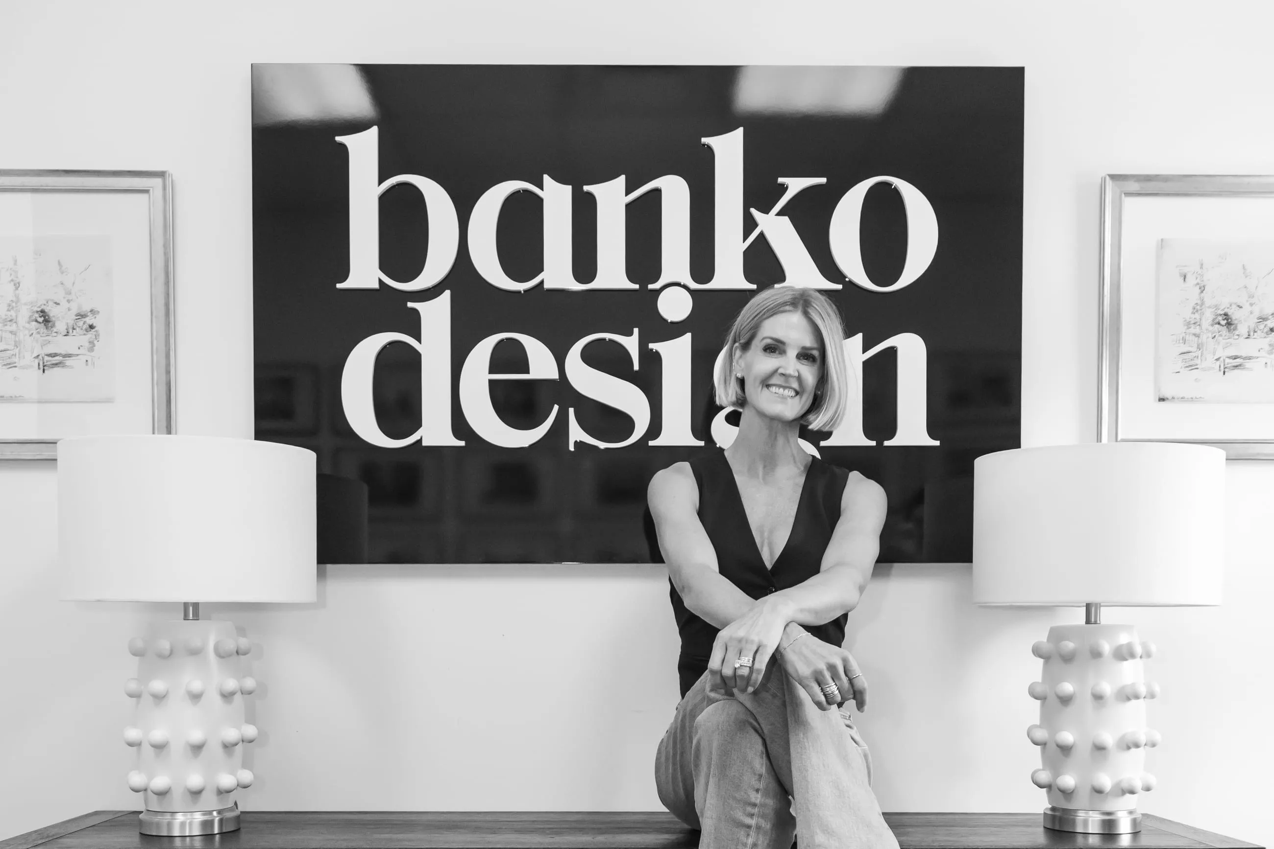 Melissa Banko seated on a console table in front of the studio's black Banko Design sign, between two white bobble table lamps, in black and white