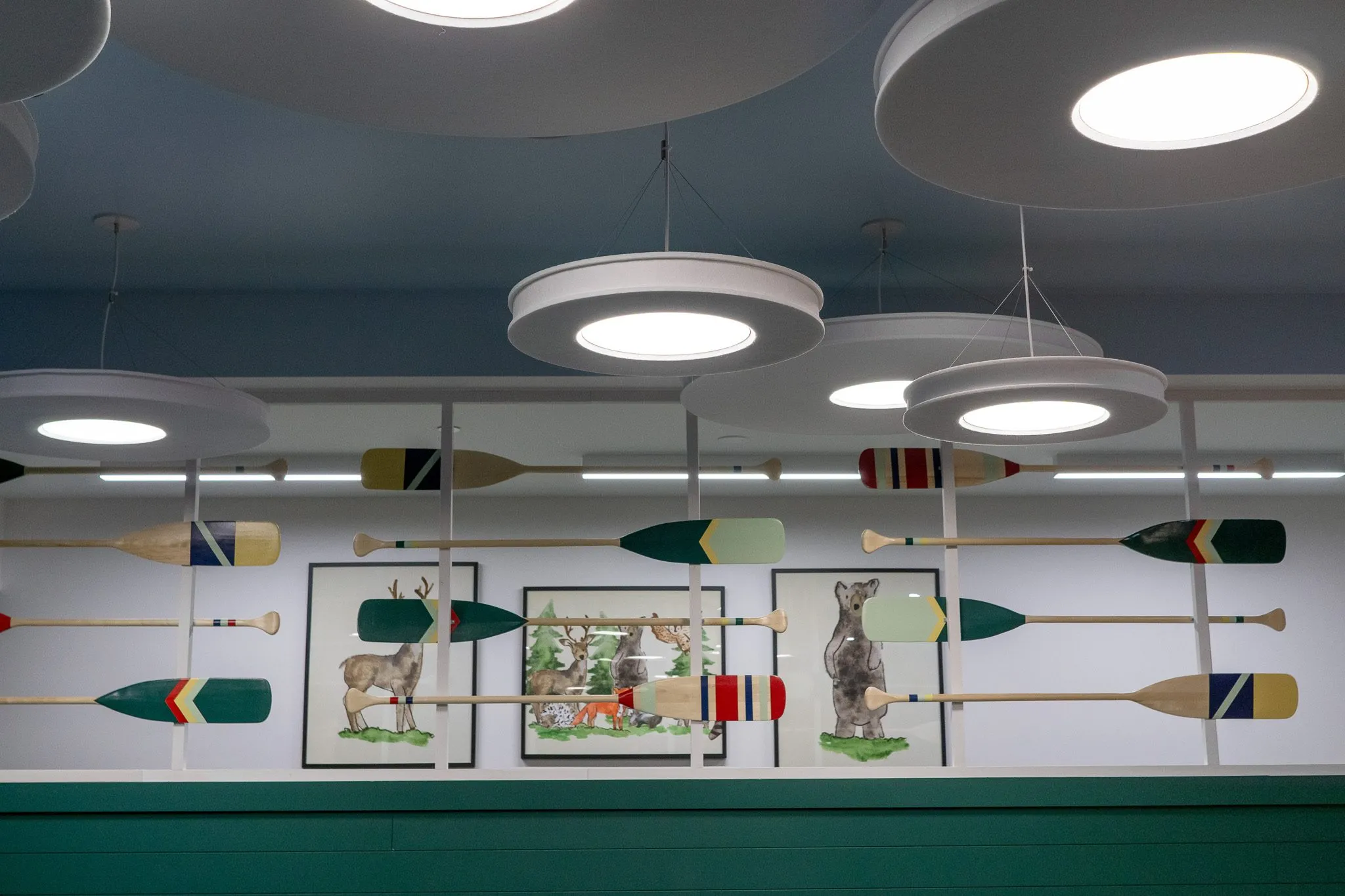 School interior detail: canoe paddles on rails across framed deer, bear and woodland prints, disc lights on the blue ceiling