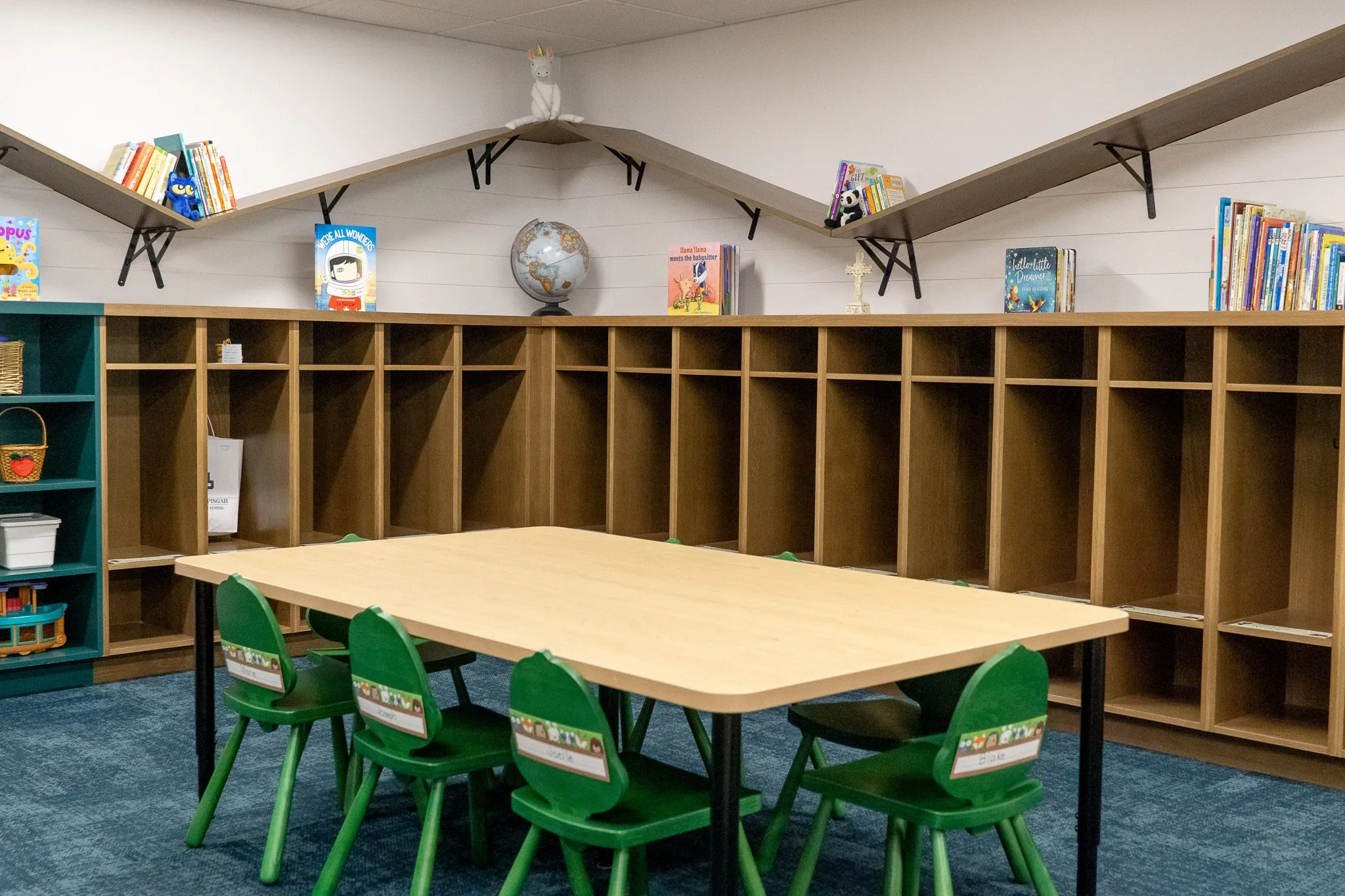 School classroom with a wood cubby wall and angled book shelves behind a table of green chairs