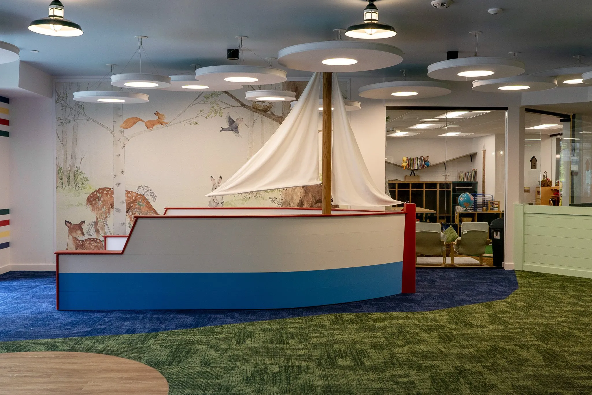 School lobby with a sailboat play structure set into the carpet, a woodland mural of deer and foxes behind it