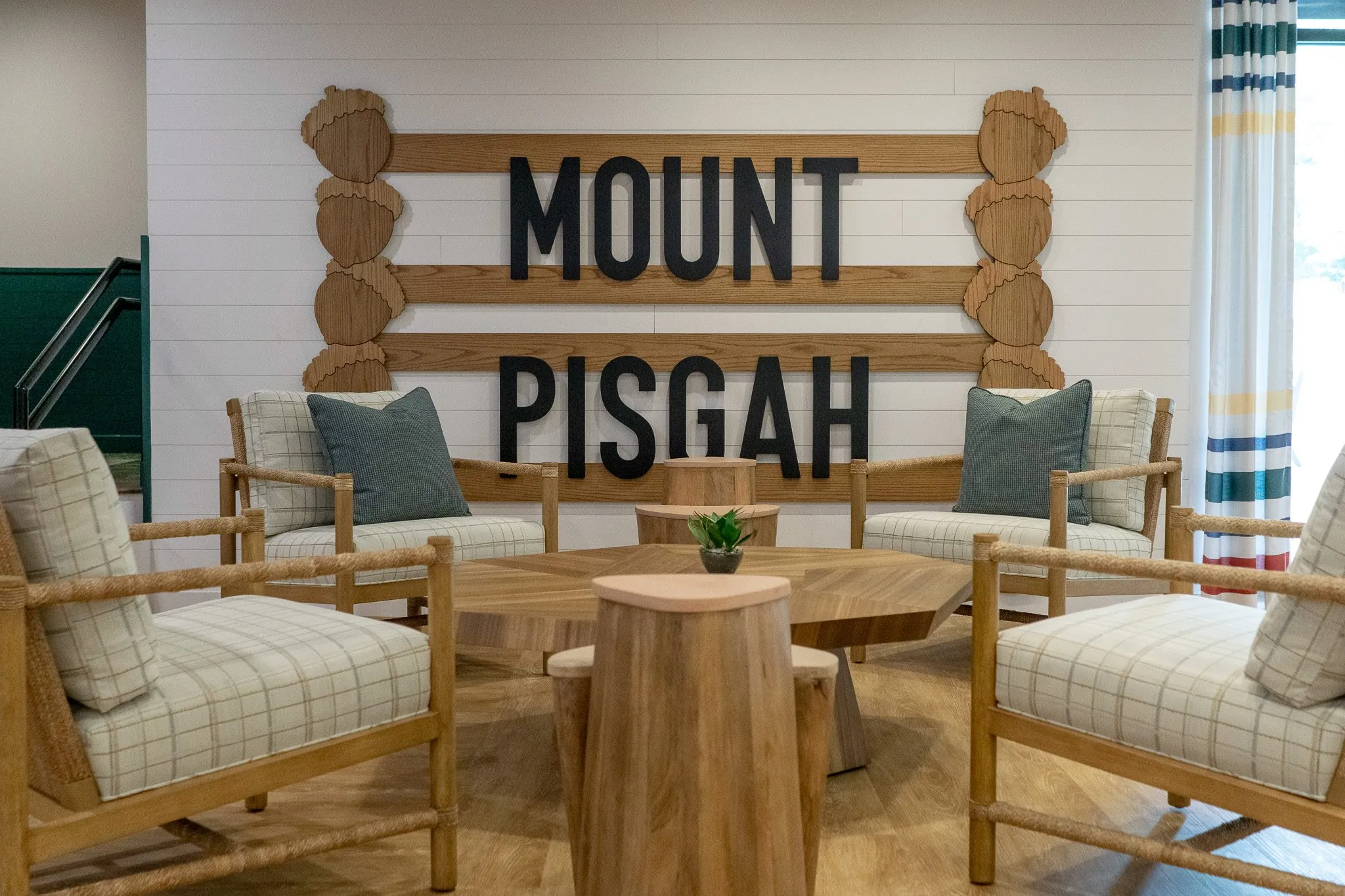 School interior detail: Mount Pisgah spelled in black letters on a log rail above plaid armchairs and a round stump table