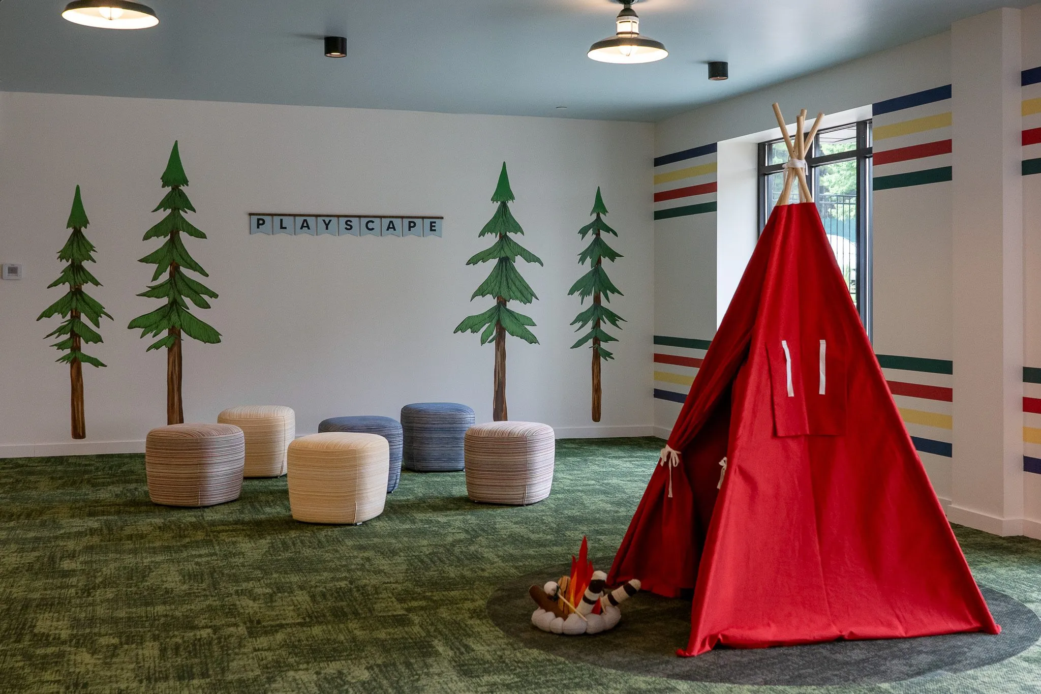 School playscape room with a red canvas teepee and a faux campfire beside woven poufs and painted pine trees