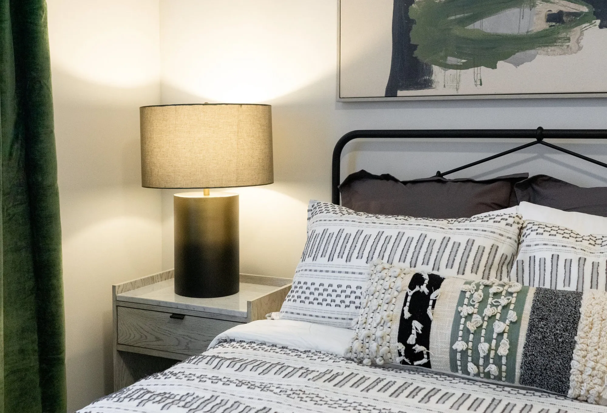 Multifamily apartment model bedroom with a black iron bed, woven bedding, a green velvet curtain, a green and navy abstract