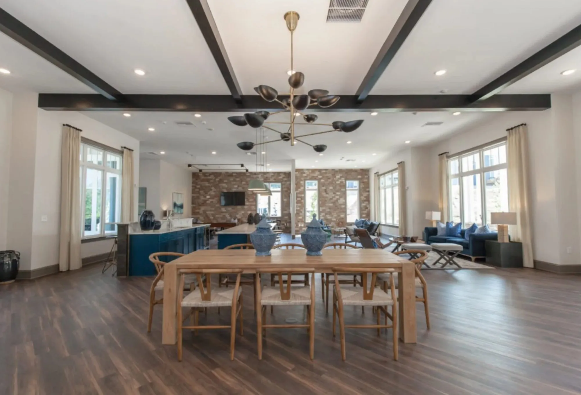 Multifamily amenity clubroom with a brass chandelier over a long communal table and wishbone chairs beside a stone-clad bar