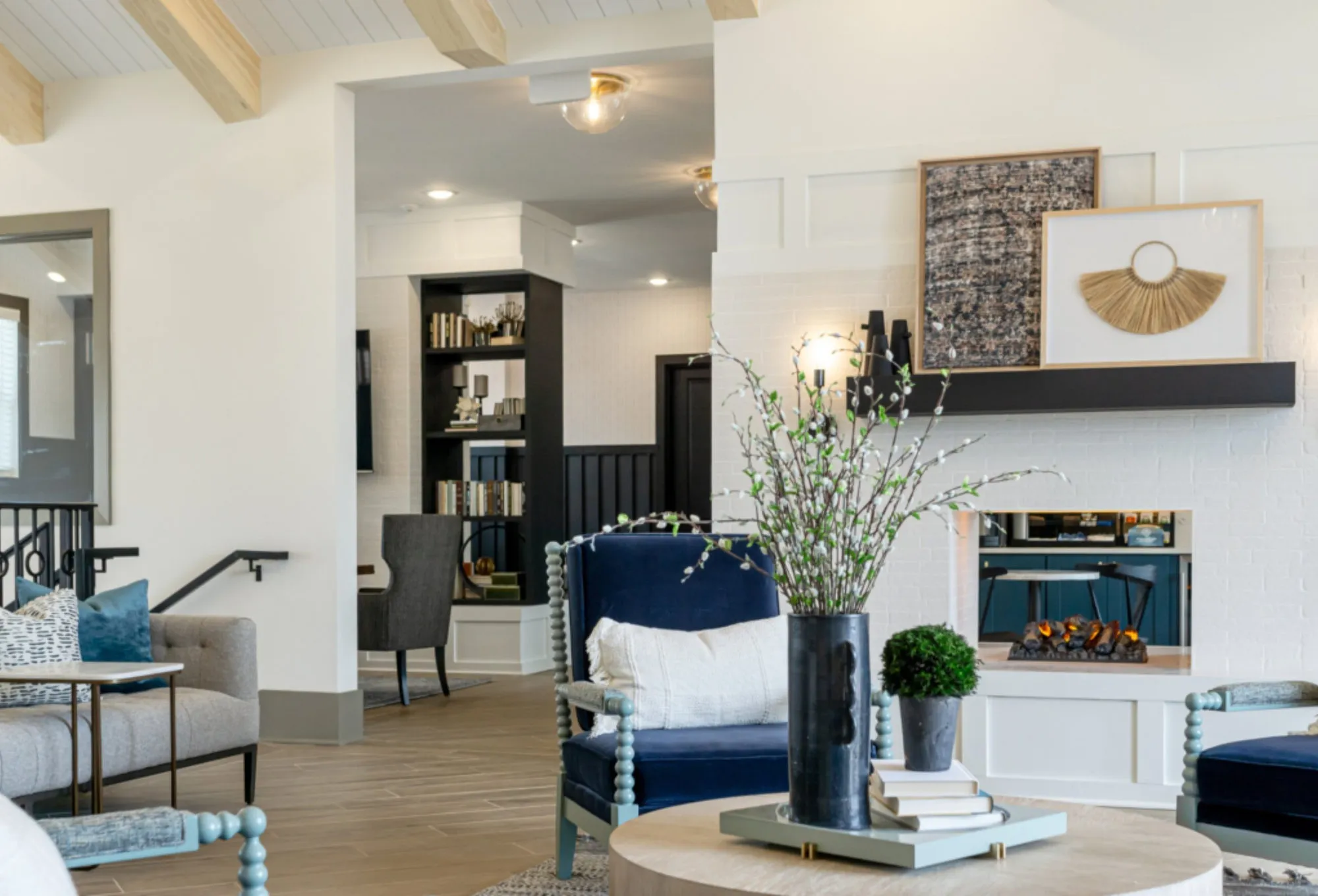 Multifamily amenity clubroom with whitewashed beams, navy spindle chairs at a white brick fireplace and a branch arrangement