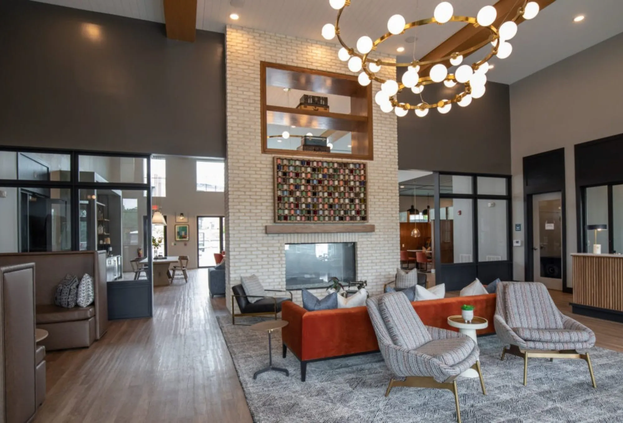 Multifamily amenity double-height leasing lobby with a brass ring chandelier over an orange sofa by a white brick fireplace