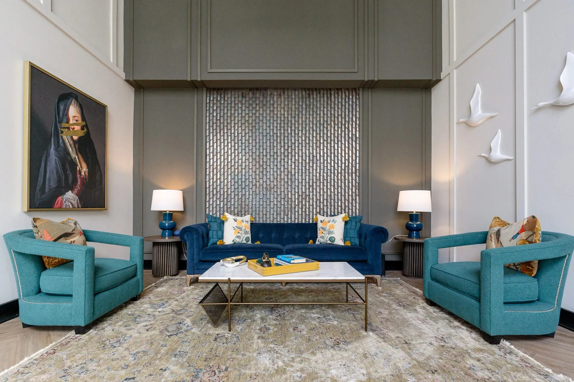 Multifamily amenity double-height lounge with teal armchairs and a navy tufted sofa below a veiled portrait, bird sculptures