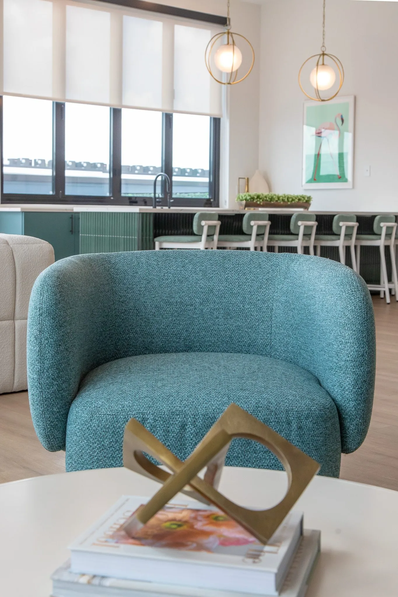 Multifamily interior detail: teal boucle barrel chair and a brass sculpture on books, kitchen island with green stools beyond