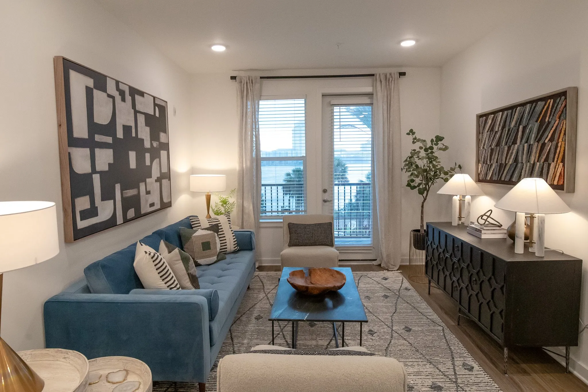 Multifamily model apartment living room with a blue velvet sofa facing balcony doors, black geometric sideboard and artworks
