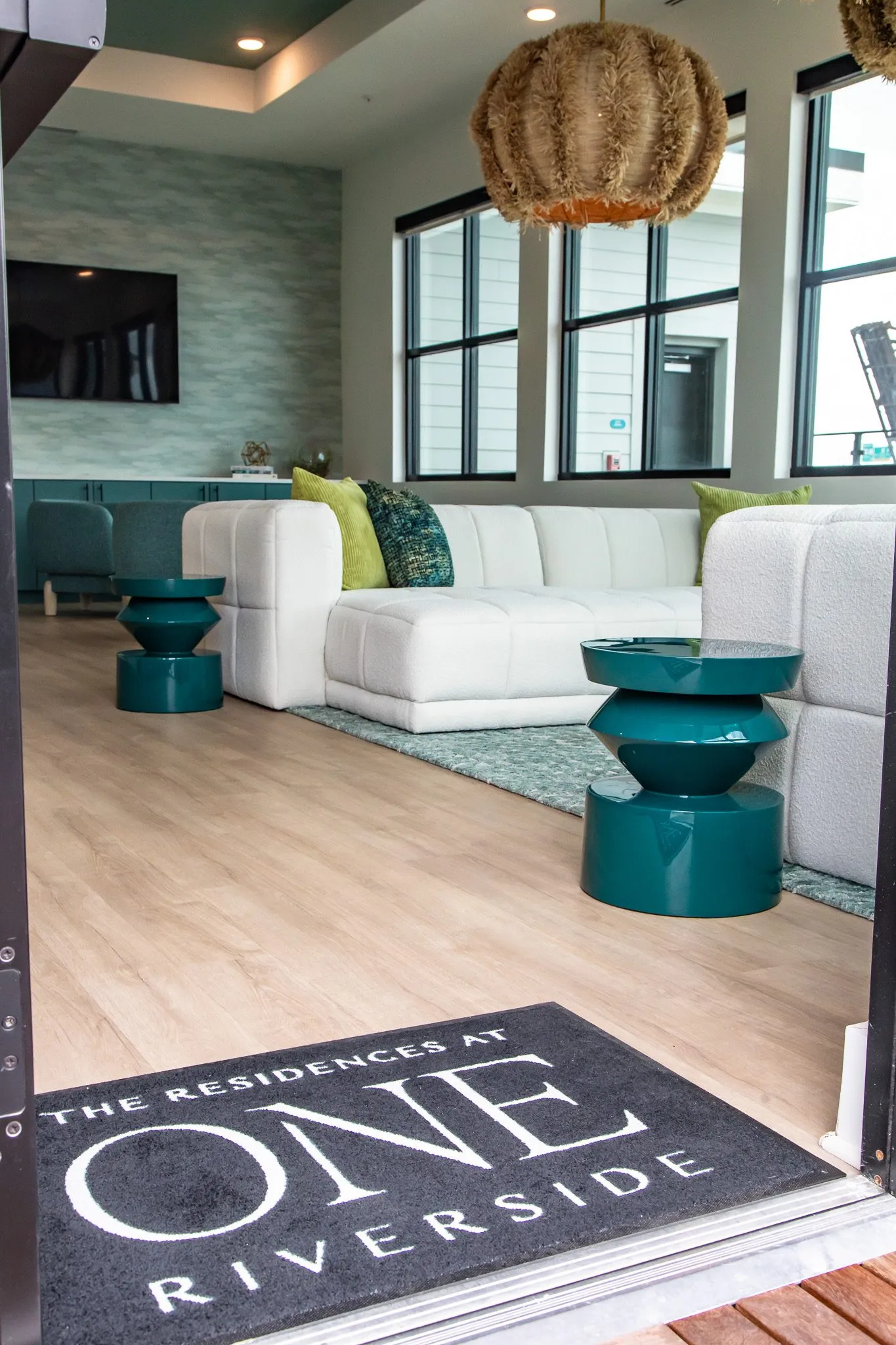 Multifamily amenity clubroom seen from the doorway over a logo entry mat, white sectional and teal side tables beyond