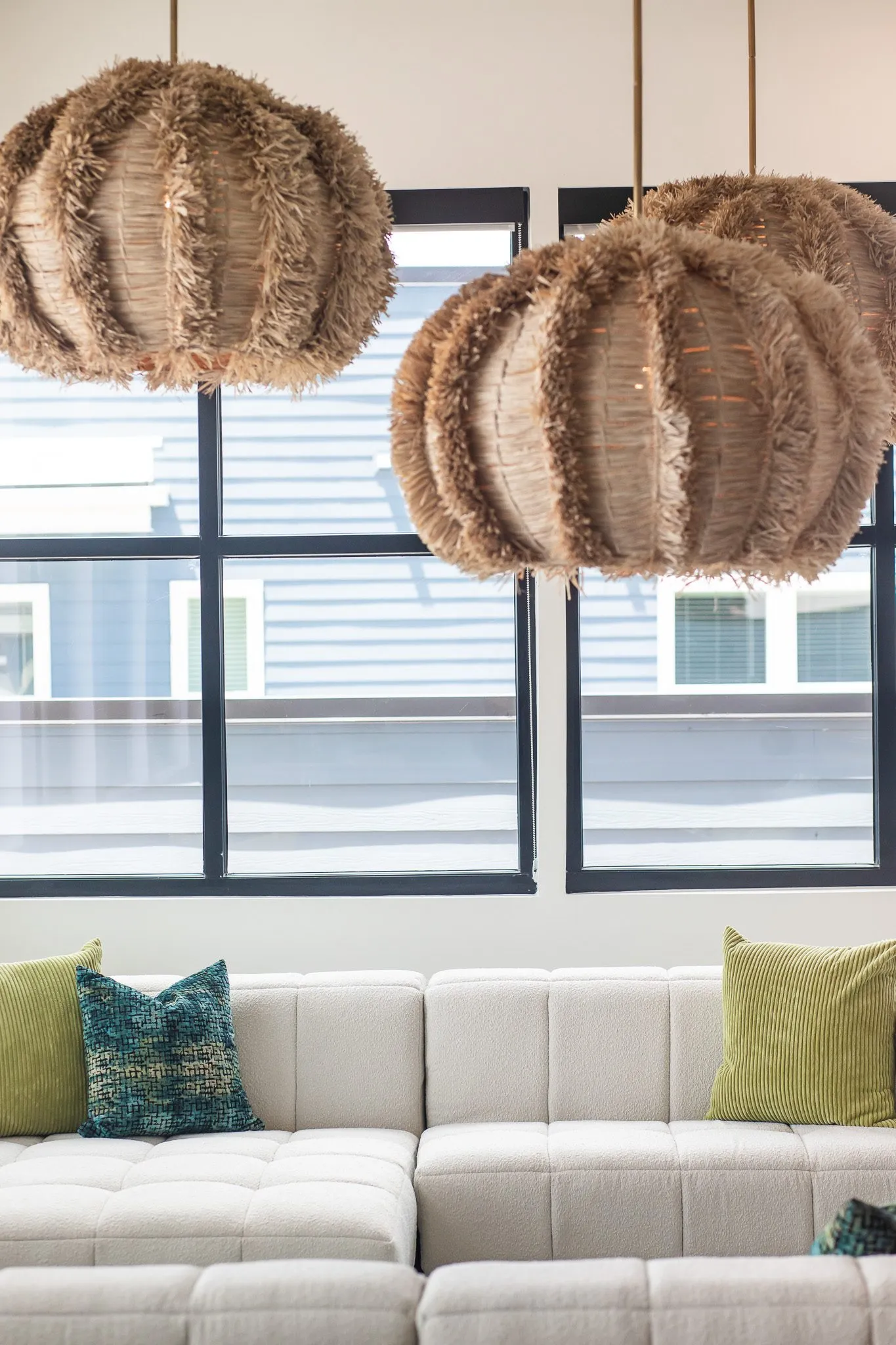 Multifamily interior detail: fringed rattan pendants above a white tufted sectional with green and teal pillows by windows