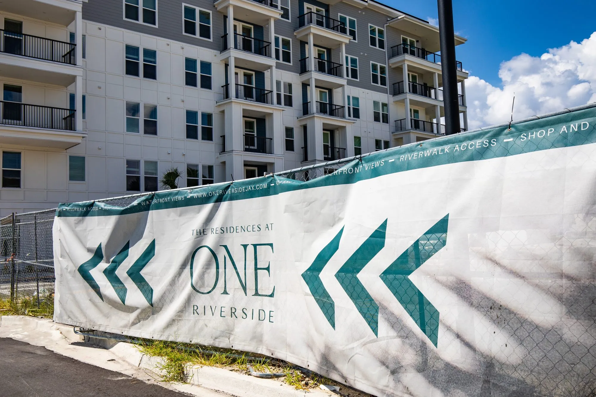 Multifamily community under construction: fence wrapped in a white banner with green chevrons beside the new mid-rise