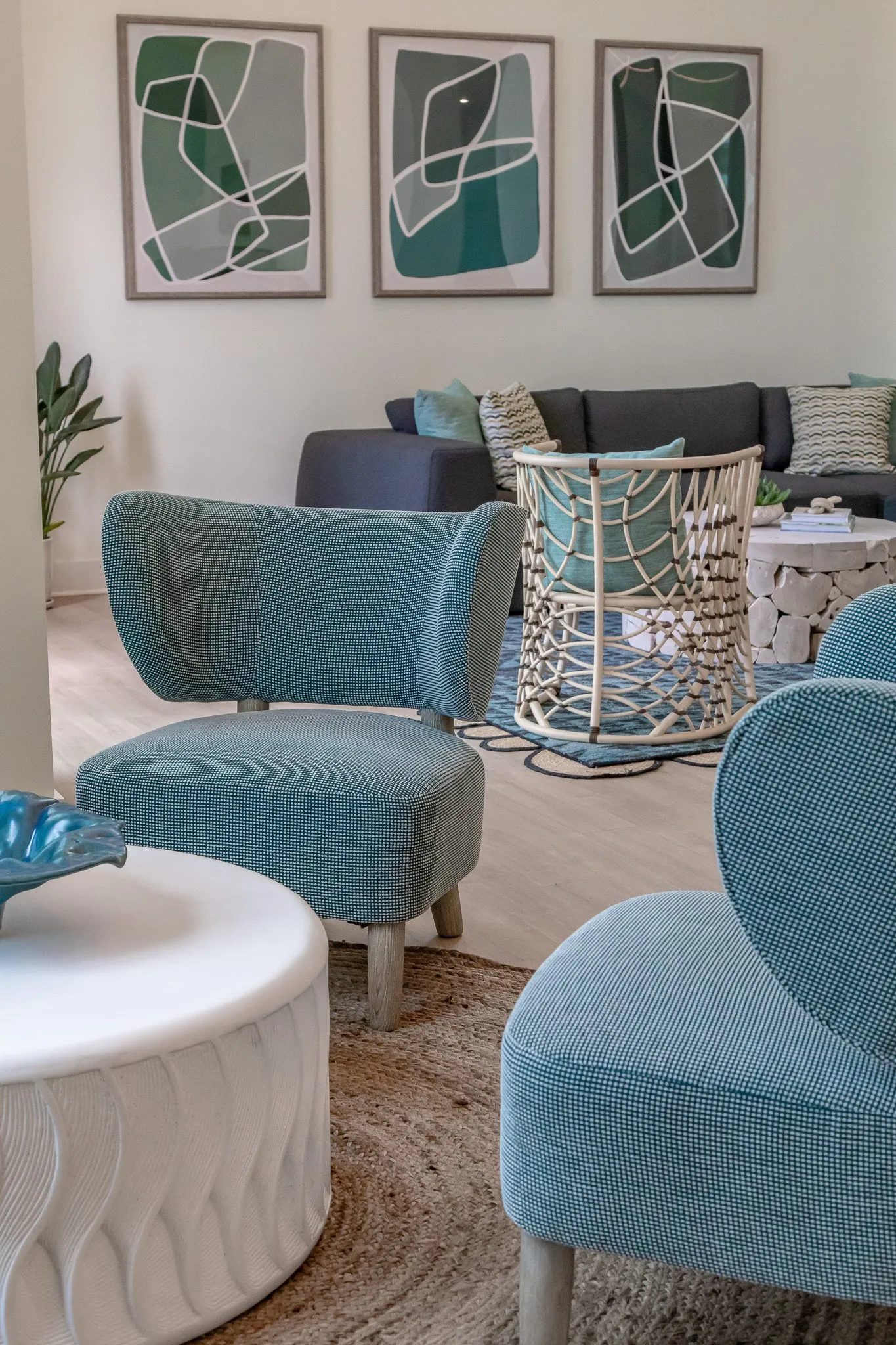 Multifamily amenity lounge with teal chairs around a white table, a grey sofa and three framed green abstract prints beyond