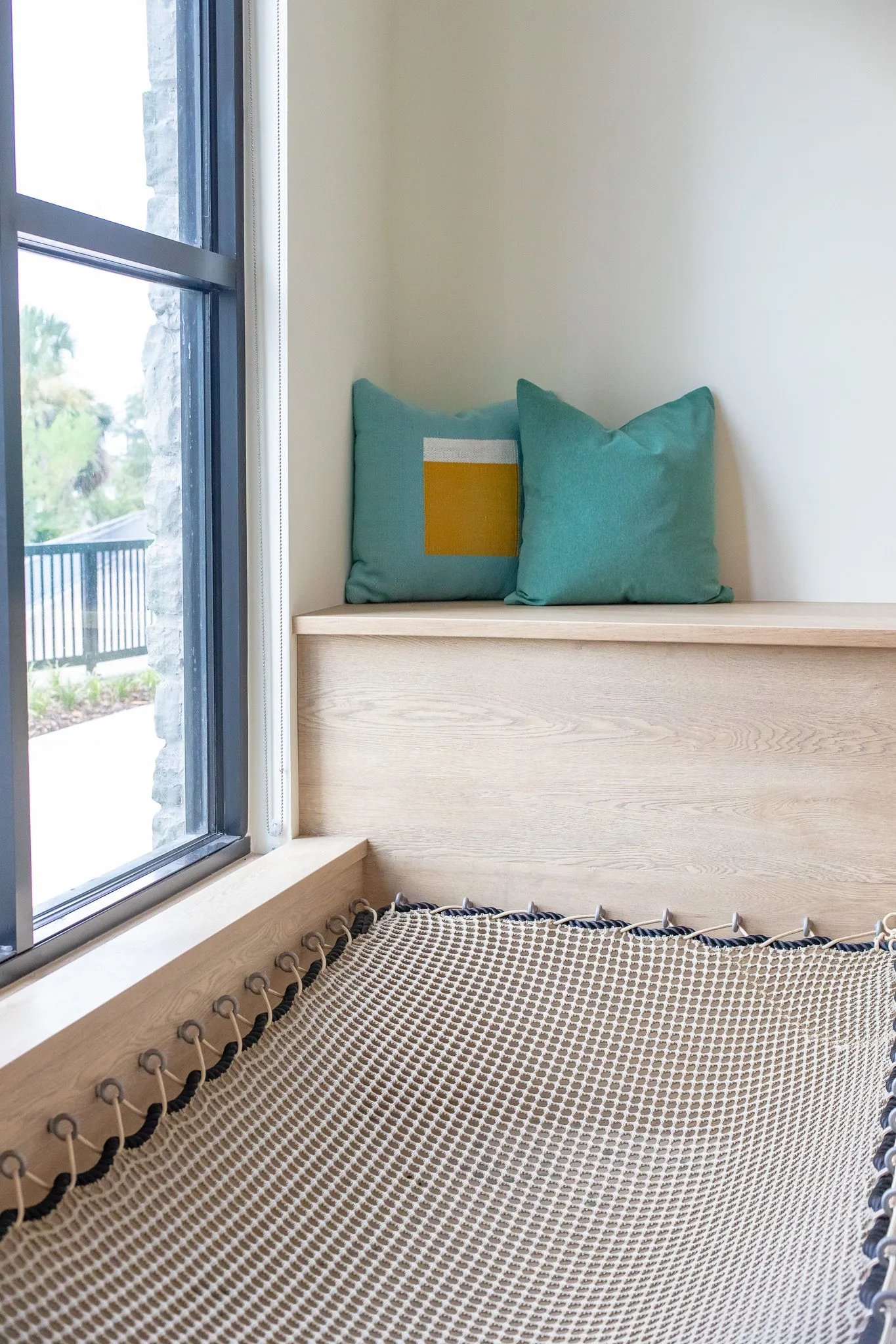 Multifamily interior detail: window seat with a rope net hammock below a wood bench and teal pillows