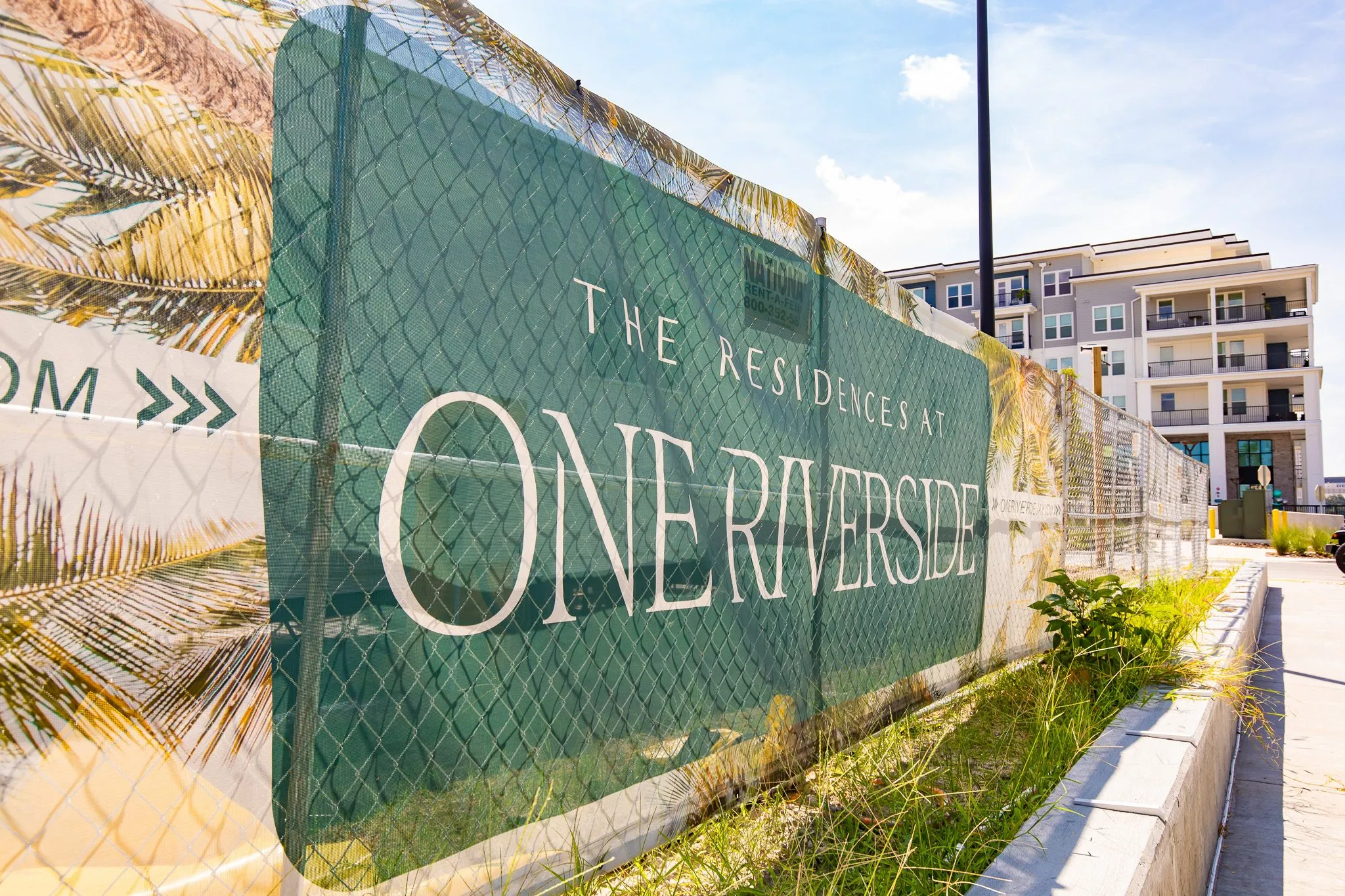 Multifamily community under construction: green fence banner with large white lettering along a curb, the new mid-rise beyond