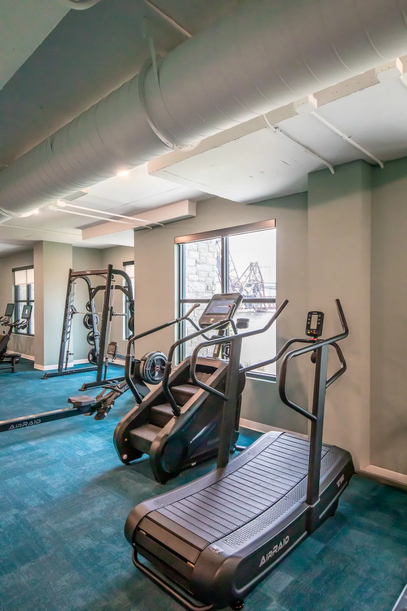 Multifamily amenity fitness center with a curved treadmill and stair climber on teal carpet under exposed ductwork