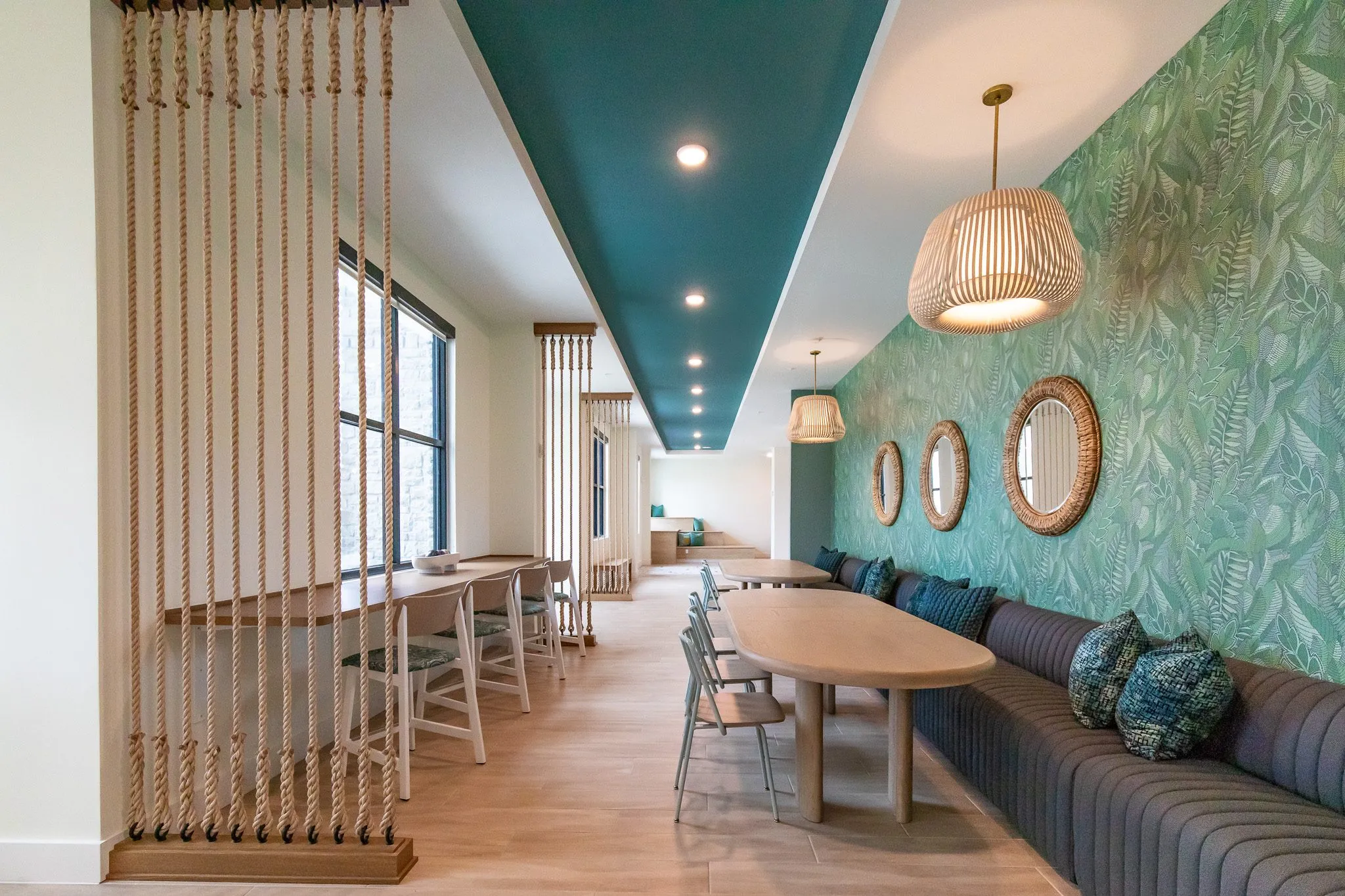 Dining lounge with a rope screen, a grey banquette along a leaf print wall hung with round rattan mirrors, and a woven pendant