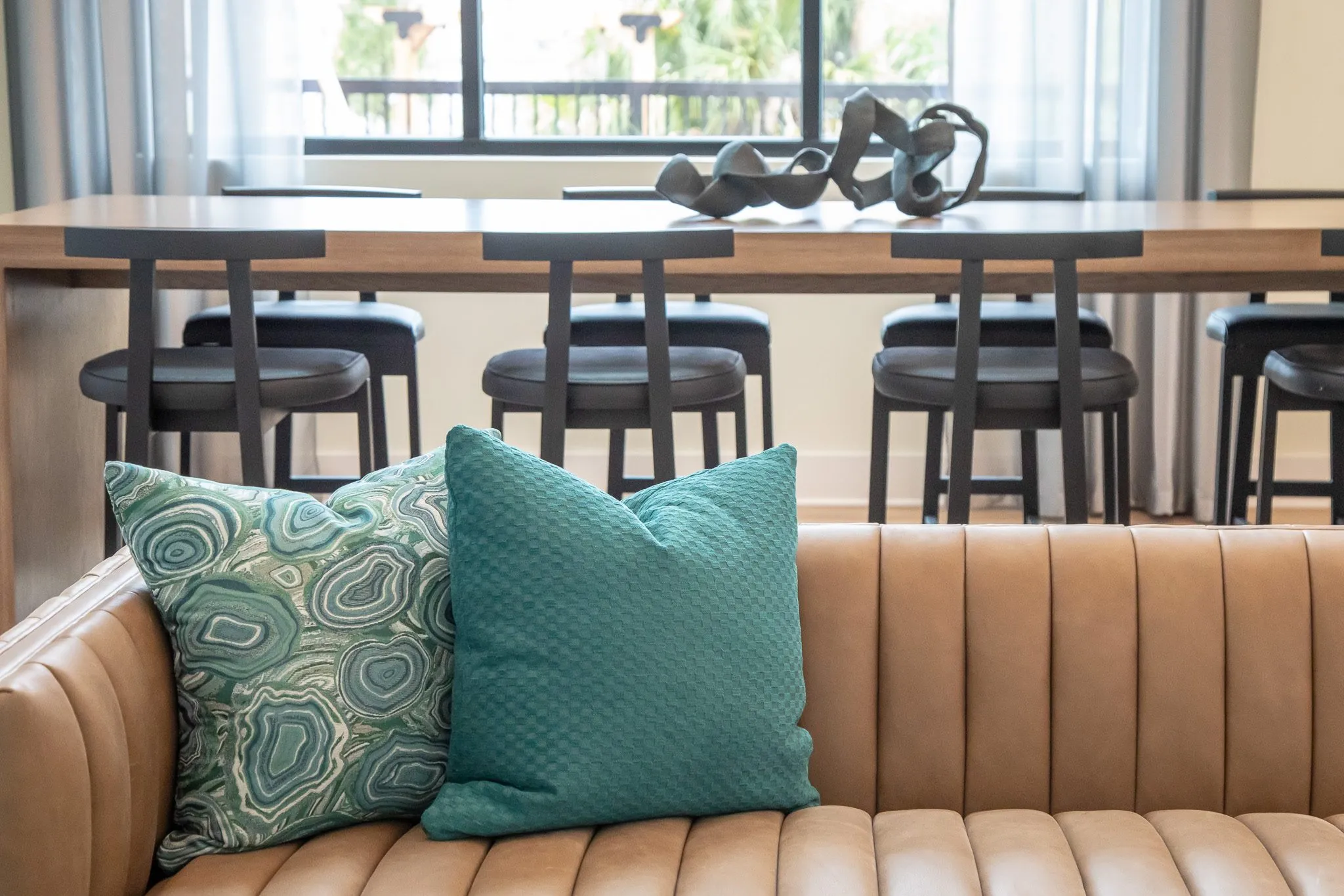 Multifamily interior detail: tan channel tufted banquette with agate and teal pillows before a communal table with stools