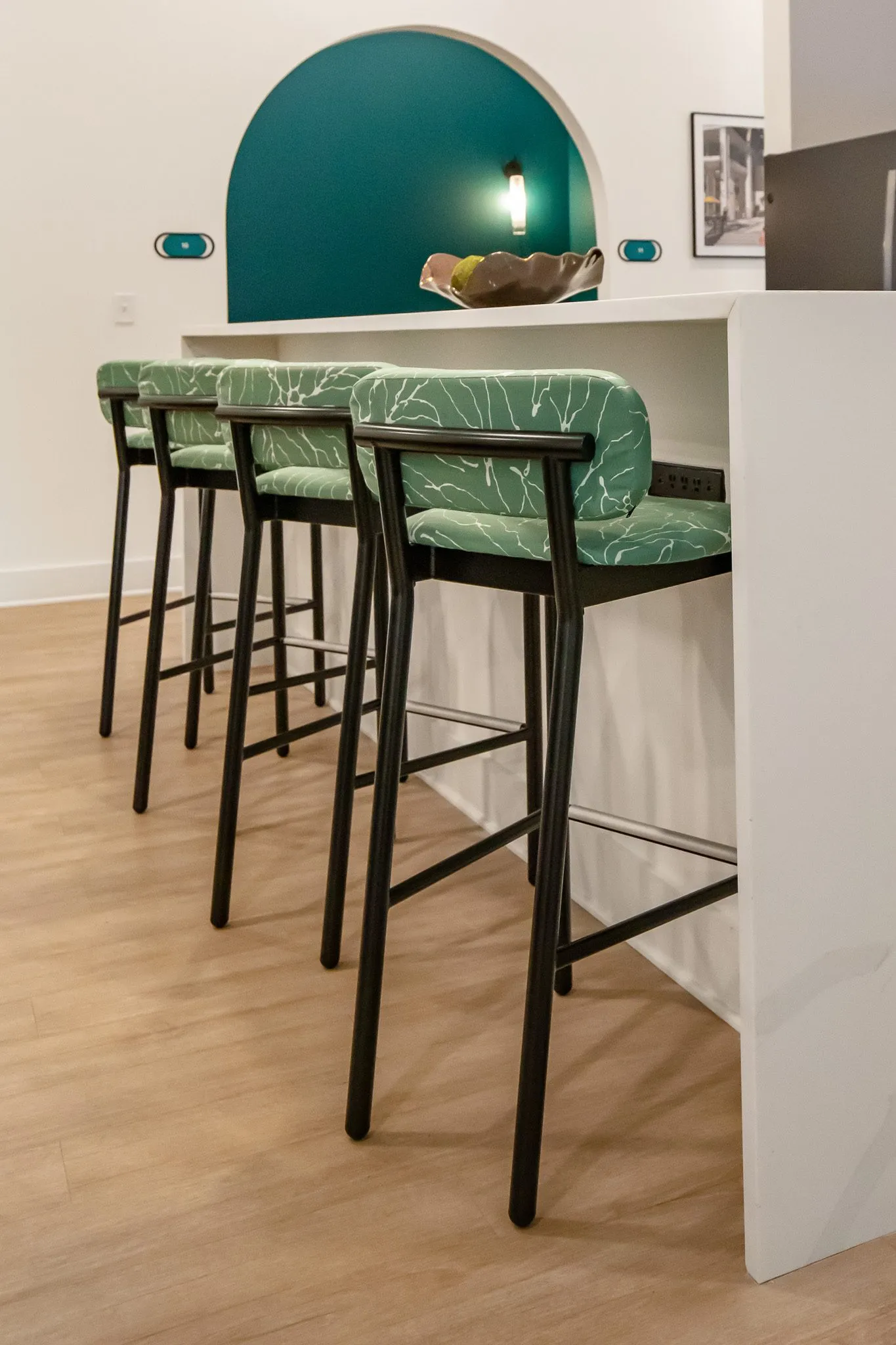 Multifamily interior detail: green leaf print barstools with black frames at a white counter below a teal arched niche